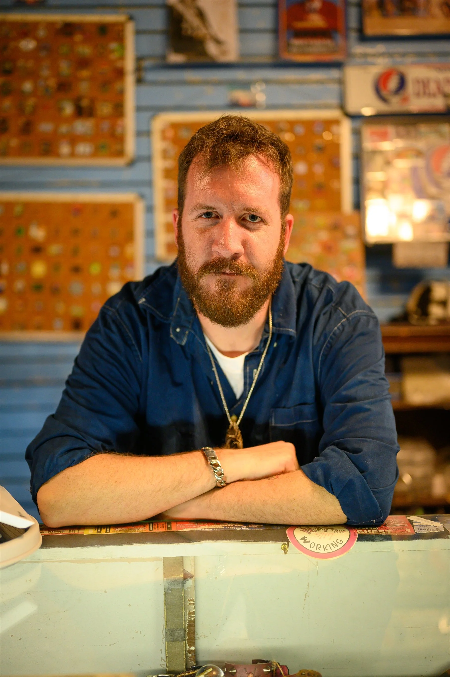 A man with a beard wearing a blue shirt stands behind a counter with glass display in a store. The background features a collection of pins or badges in wooden frames hanging on a blue wall.