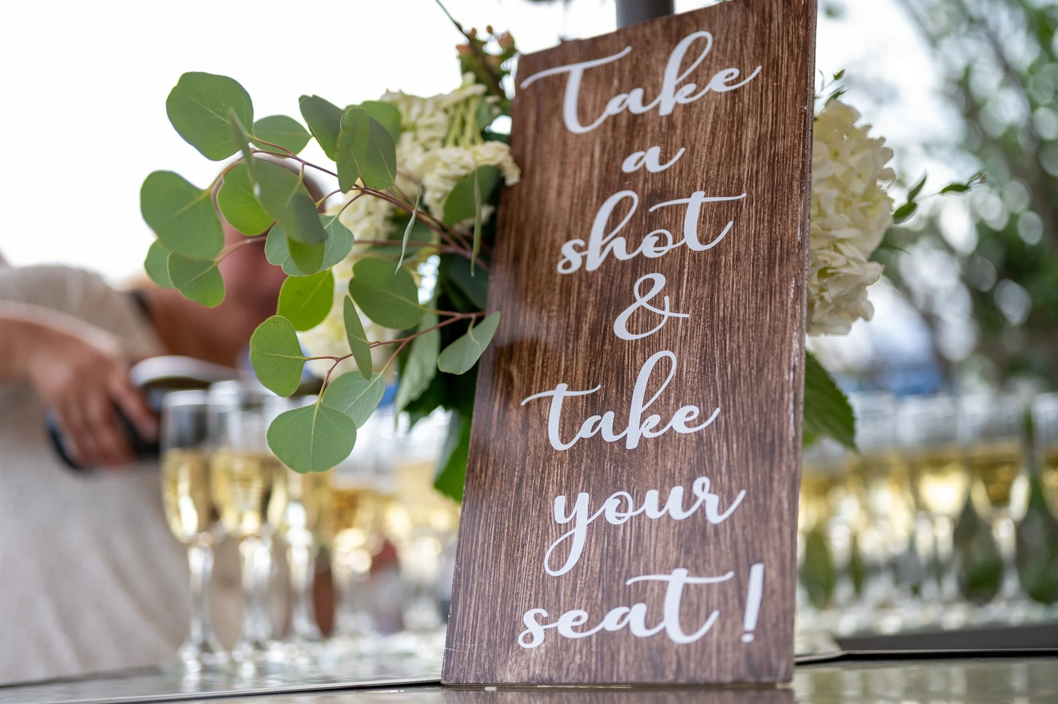 Rustic wooden sign with "Take a shot & take your seat!" near wine glasses and floral decor