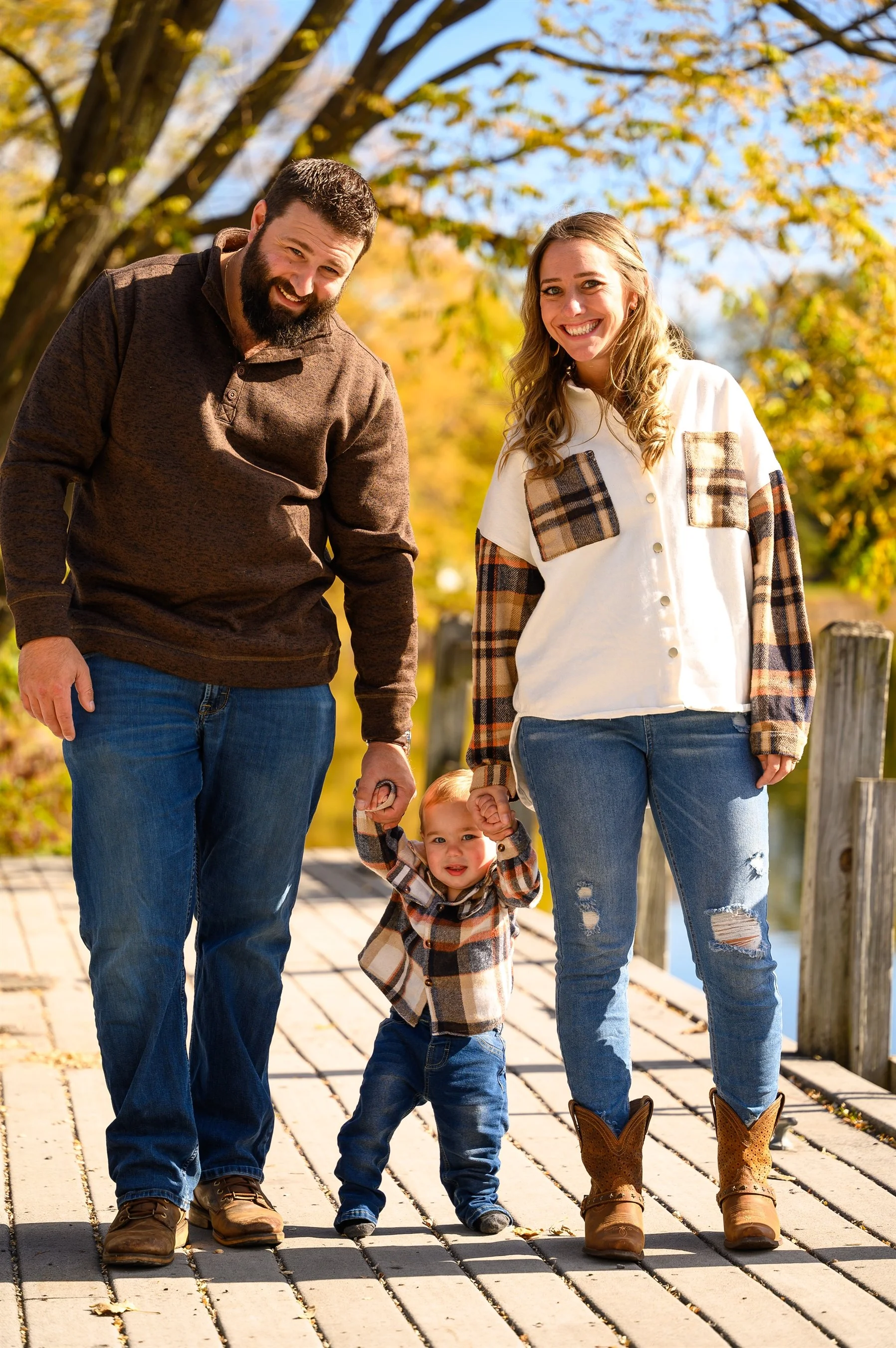 A smiling man and woman holding hands with a toddler on a wooden walkway. All are wearing casual fall clothing, with autumn trees in the background.