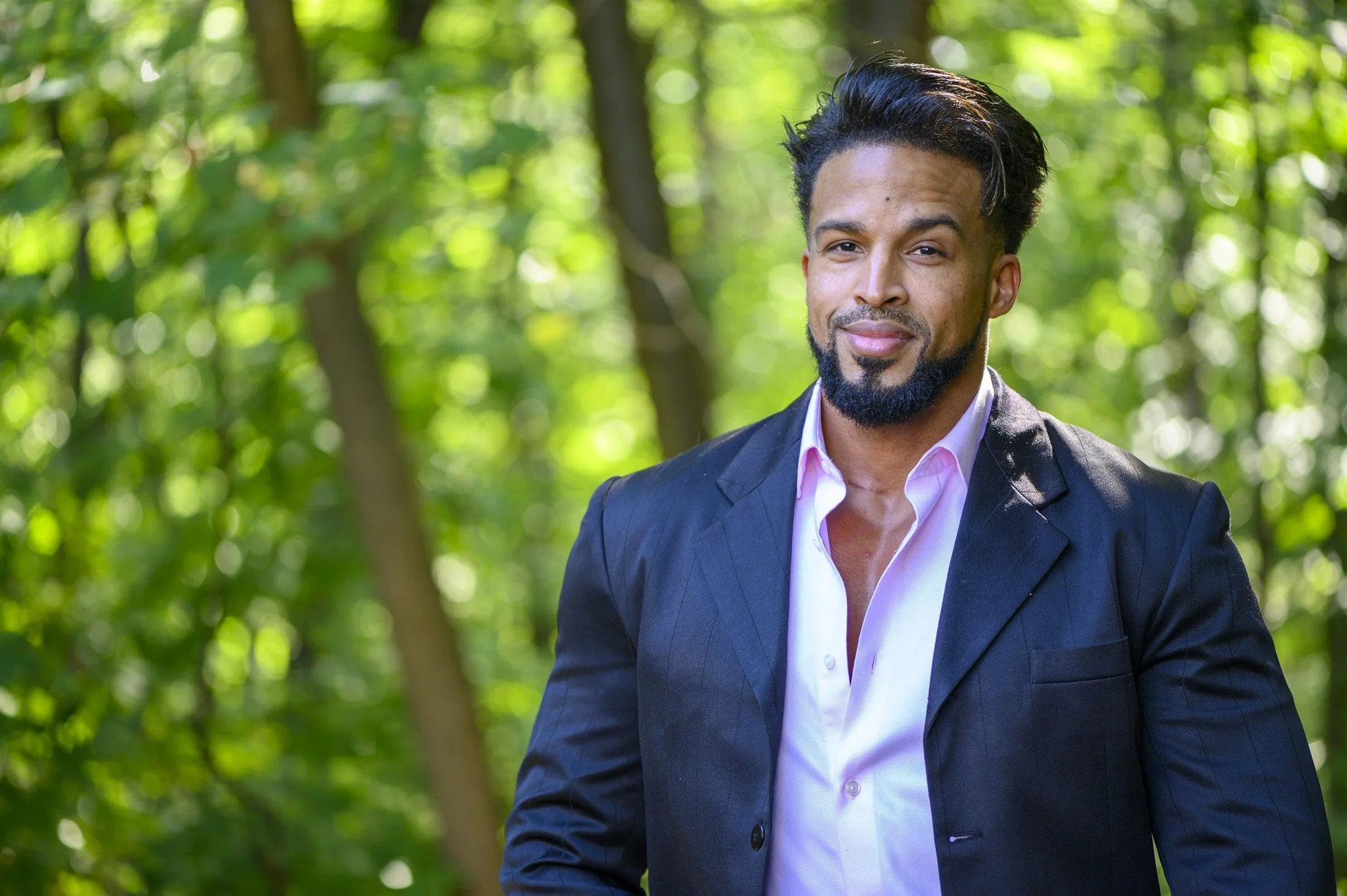 Man in a suit outdoors with trees in the background, wearing a black blazer and pink shirt, smiling.
