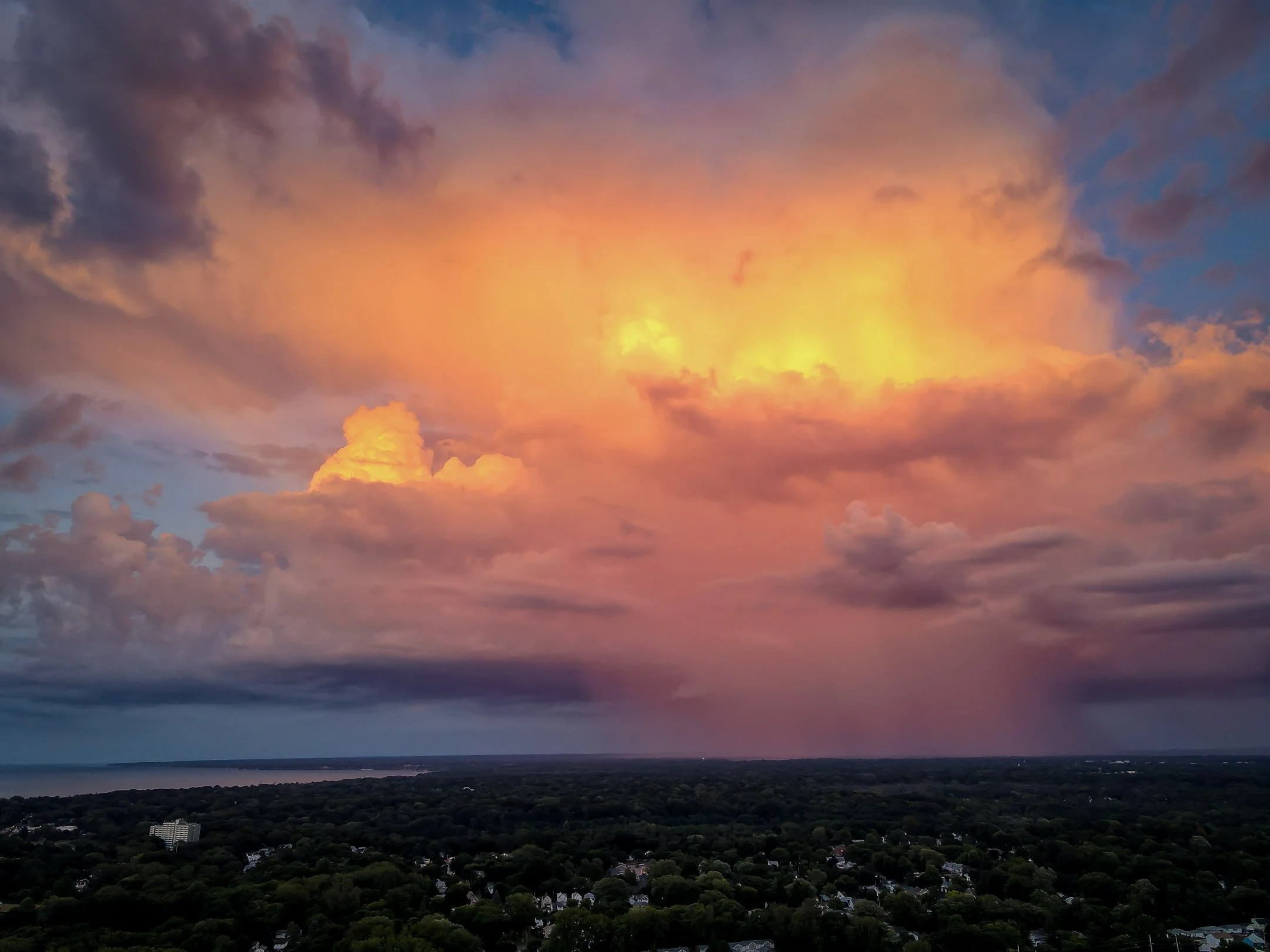 Aerial view of a vibrant sunset with dramatic clouds over a landscape of trees and scattered buildings.