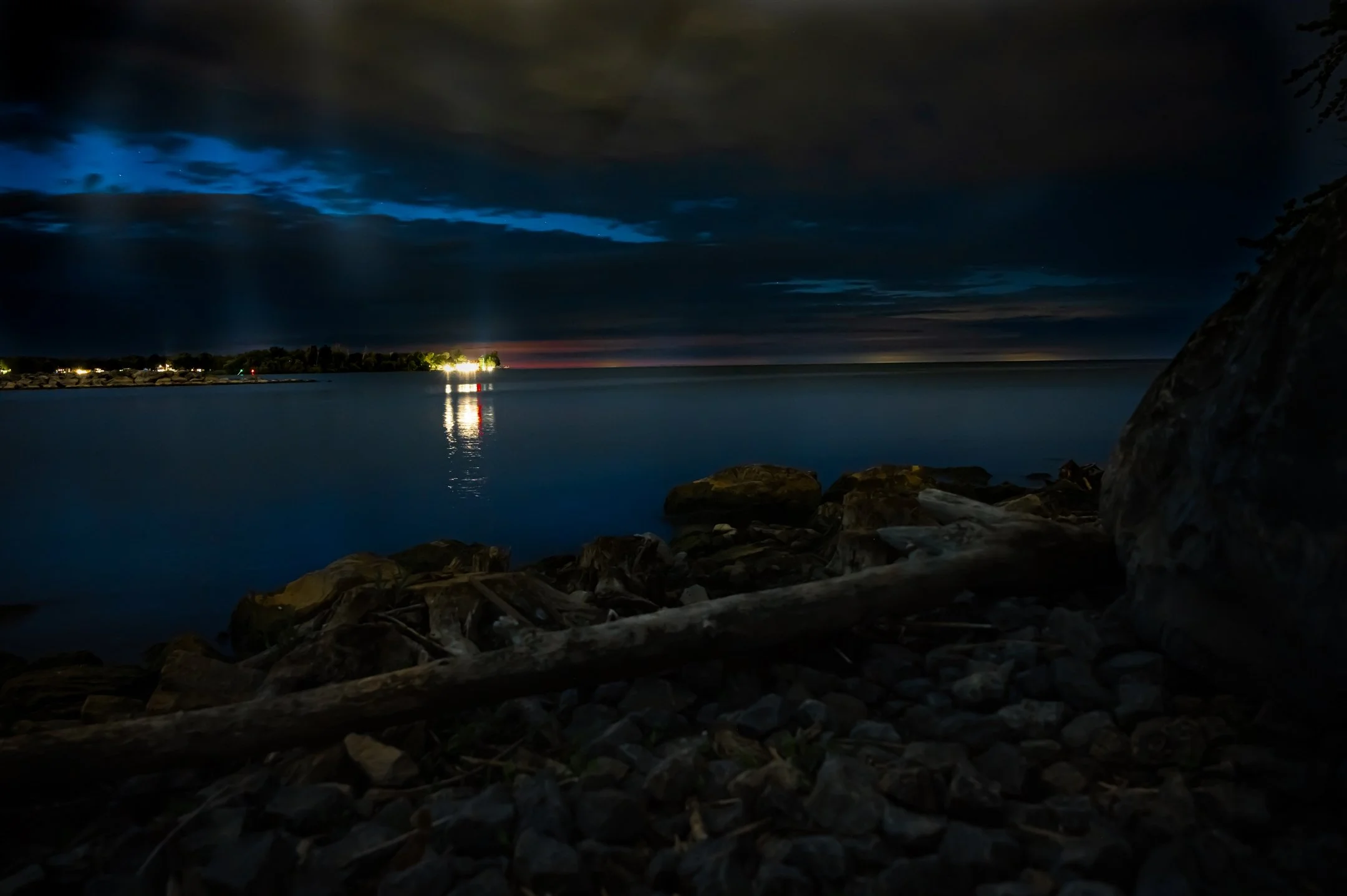 Night view of a lake with a rocky shoreline and driftwood in the foreground. Distant island with illuminated buildings reflects on the water under a cloudy sky.