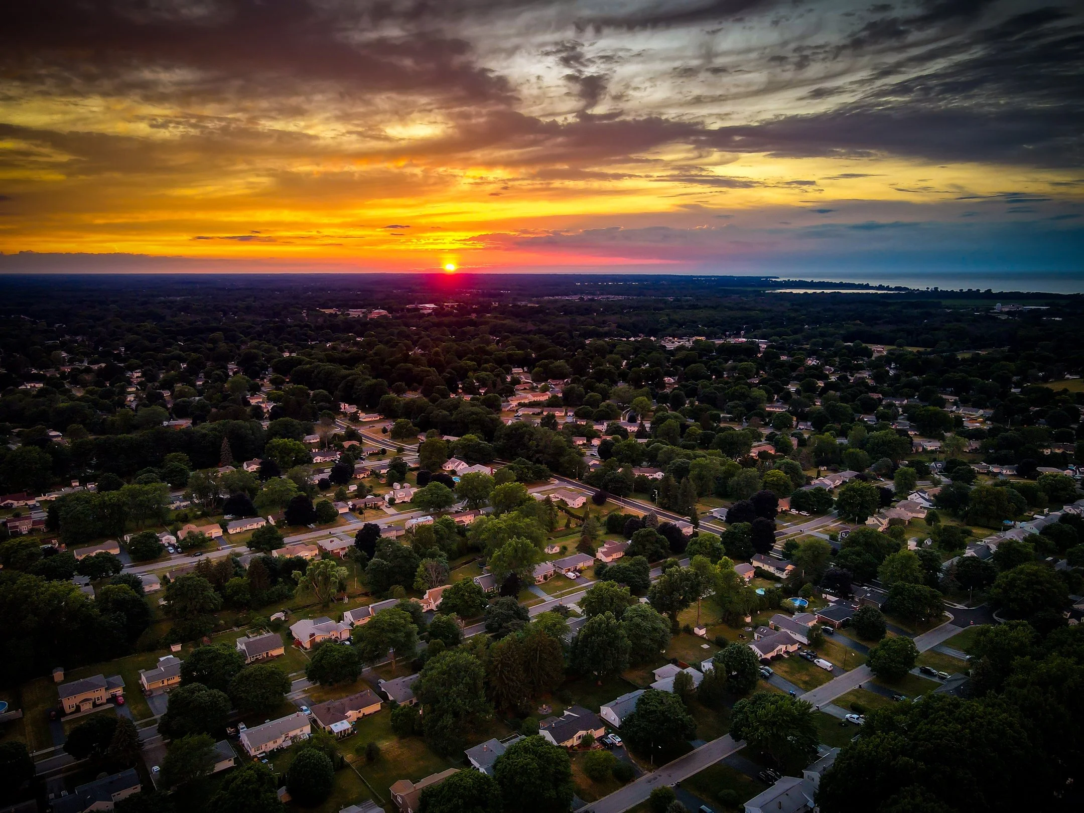 Aerial view of suburban neighborhood at sunset with a mix of houses and trees under a vibrant sky.