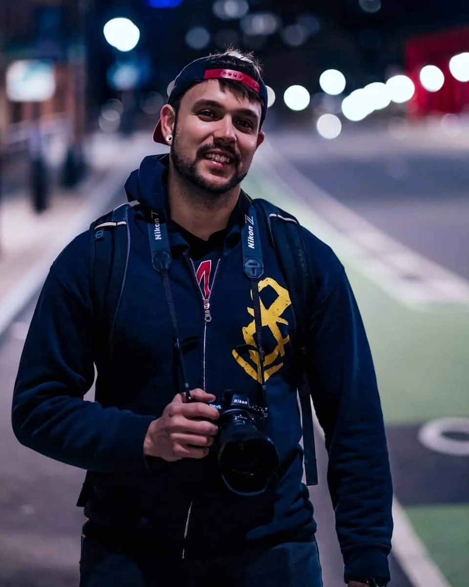 A man holding a camera on a dimly lit street at night, wearing a navy hoodie with yellow print and a backwards baseball cap.