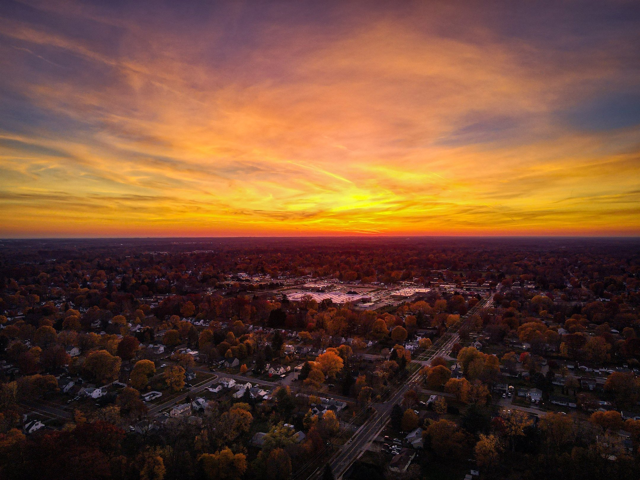 Aerial view of a suburban area with a dramatic sunset sky, showcasing vibrant orange and yellow clouds above the rooftops and streets below.
