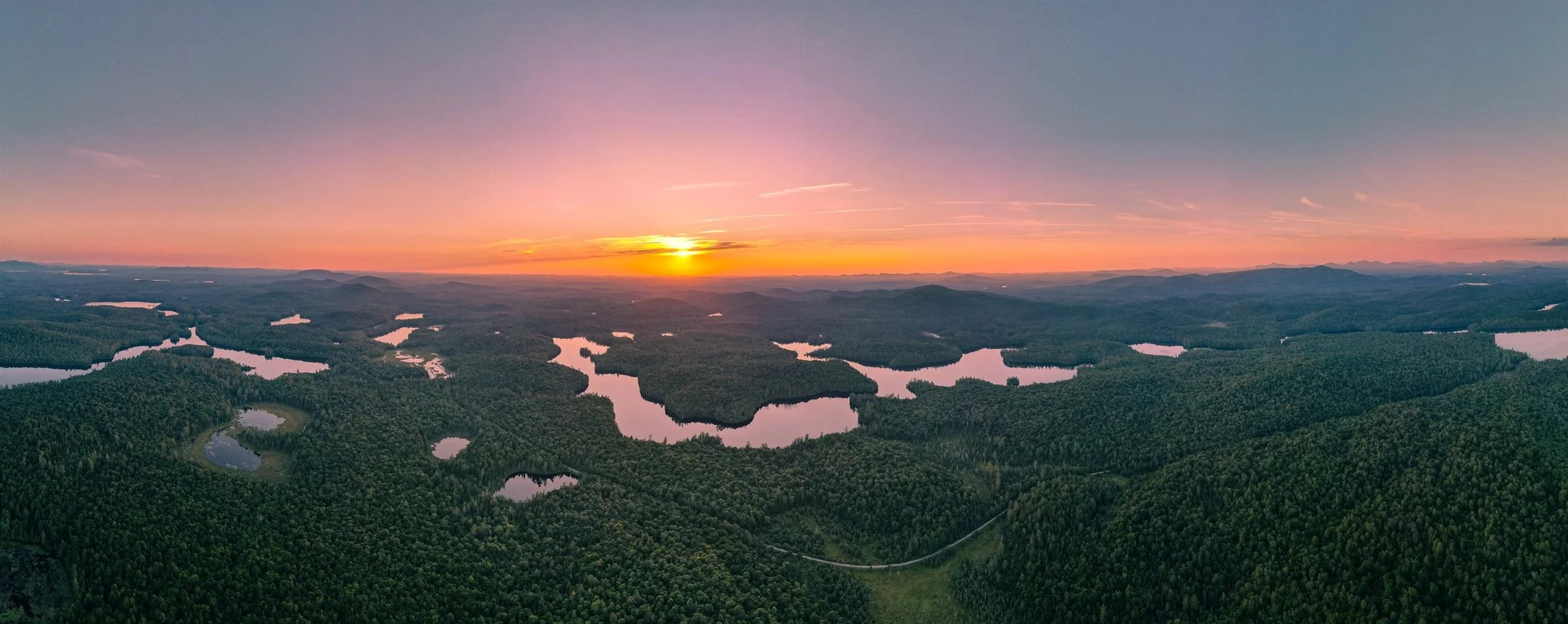 Aerial view of a forested landscape with lakes at sunset, featuring a vibrant orange and purple sky.