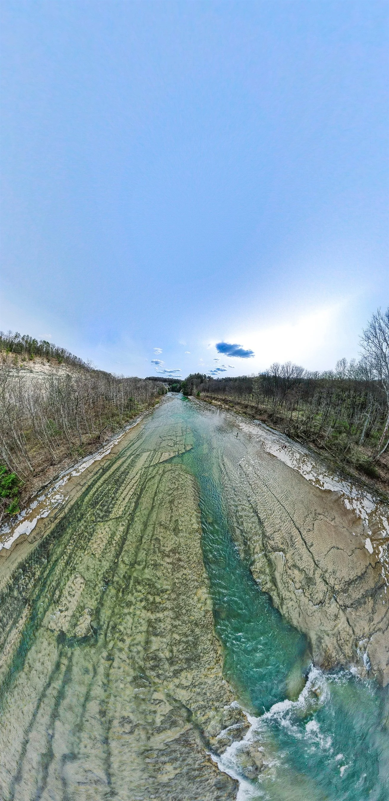 Aerial view of a clear, shallow river with rocky bottom, surrounded by bare trees and forested areas under a blue sky with some clouds.