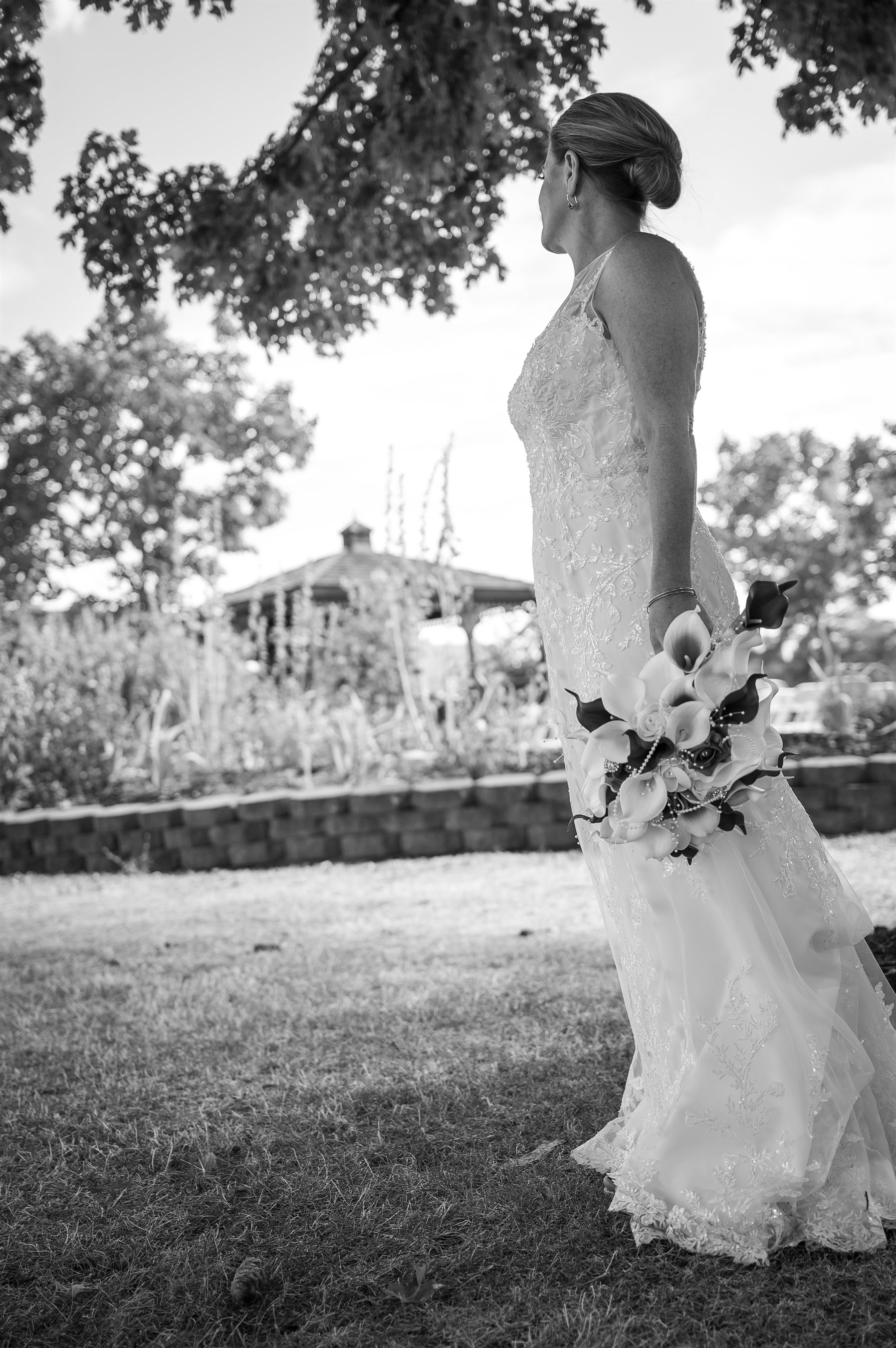 Black and white photo of a bride in a wedding dress holding a bouquet, standing outdoors under trees.