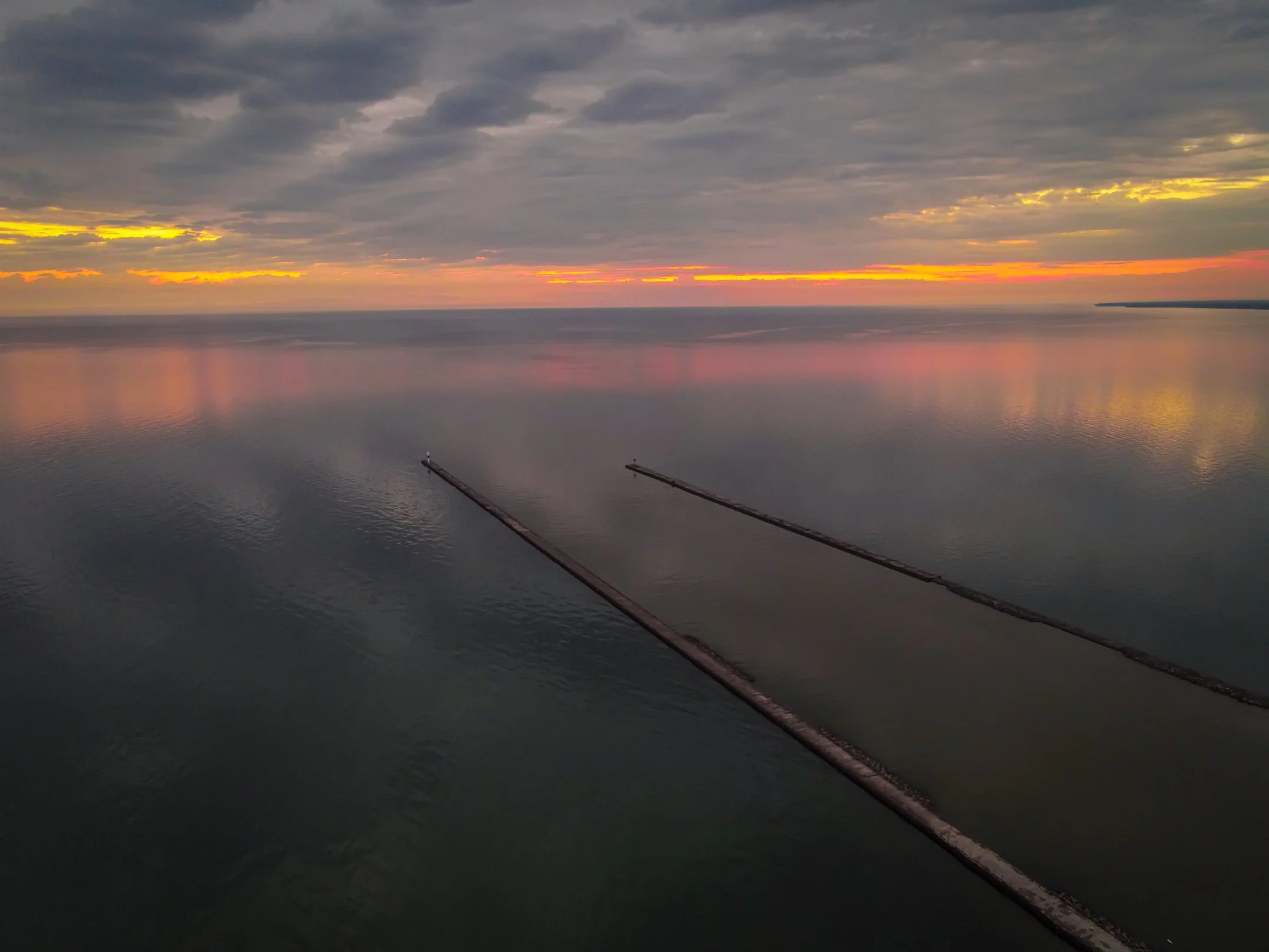 Colorful sunset over calm sea with cloudy sky and two narrow piers extending into the water.