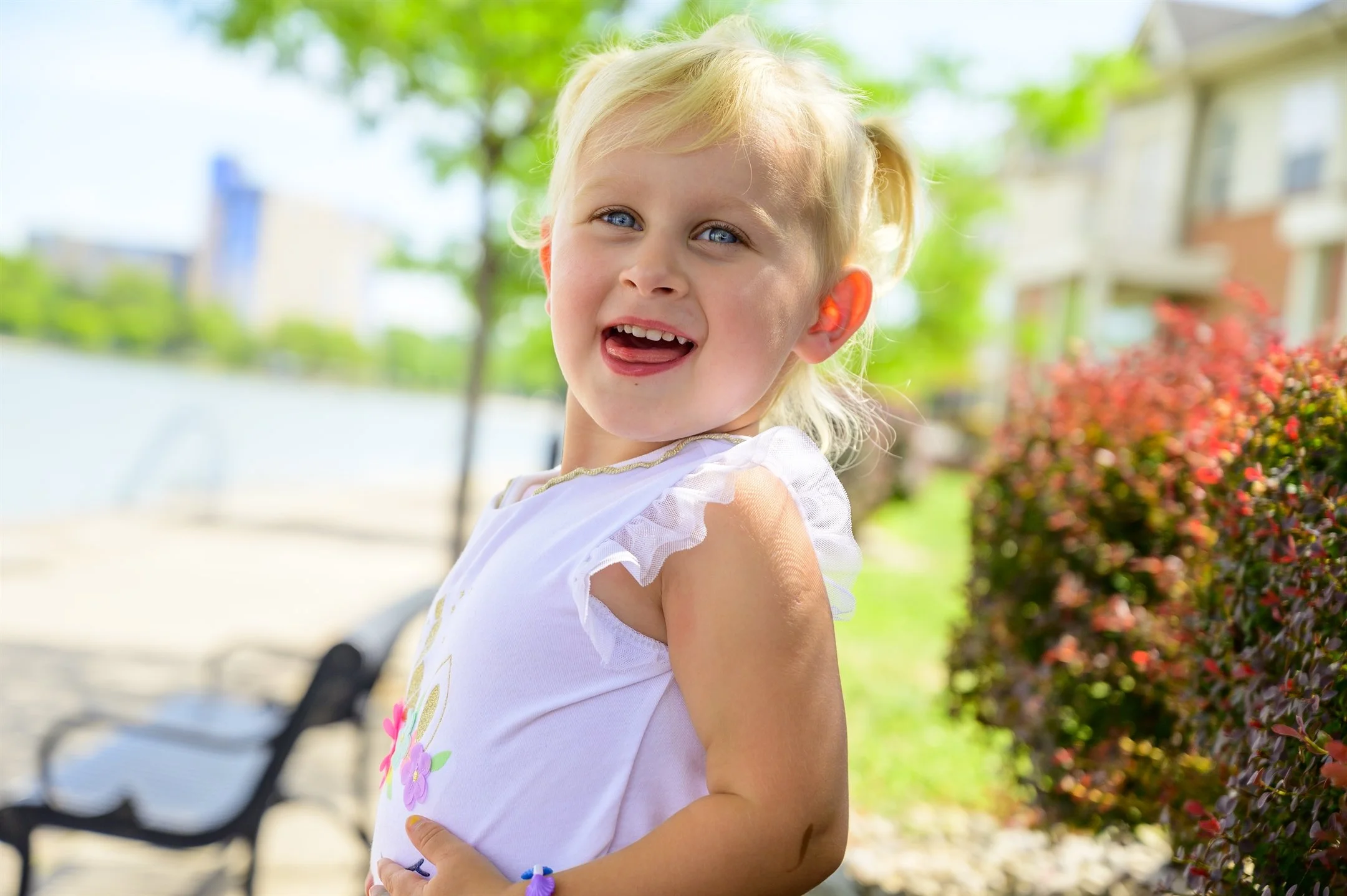 Smiling child outside near water, wearing a white dress with colorful flowers, with houses and greenery in the background.