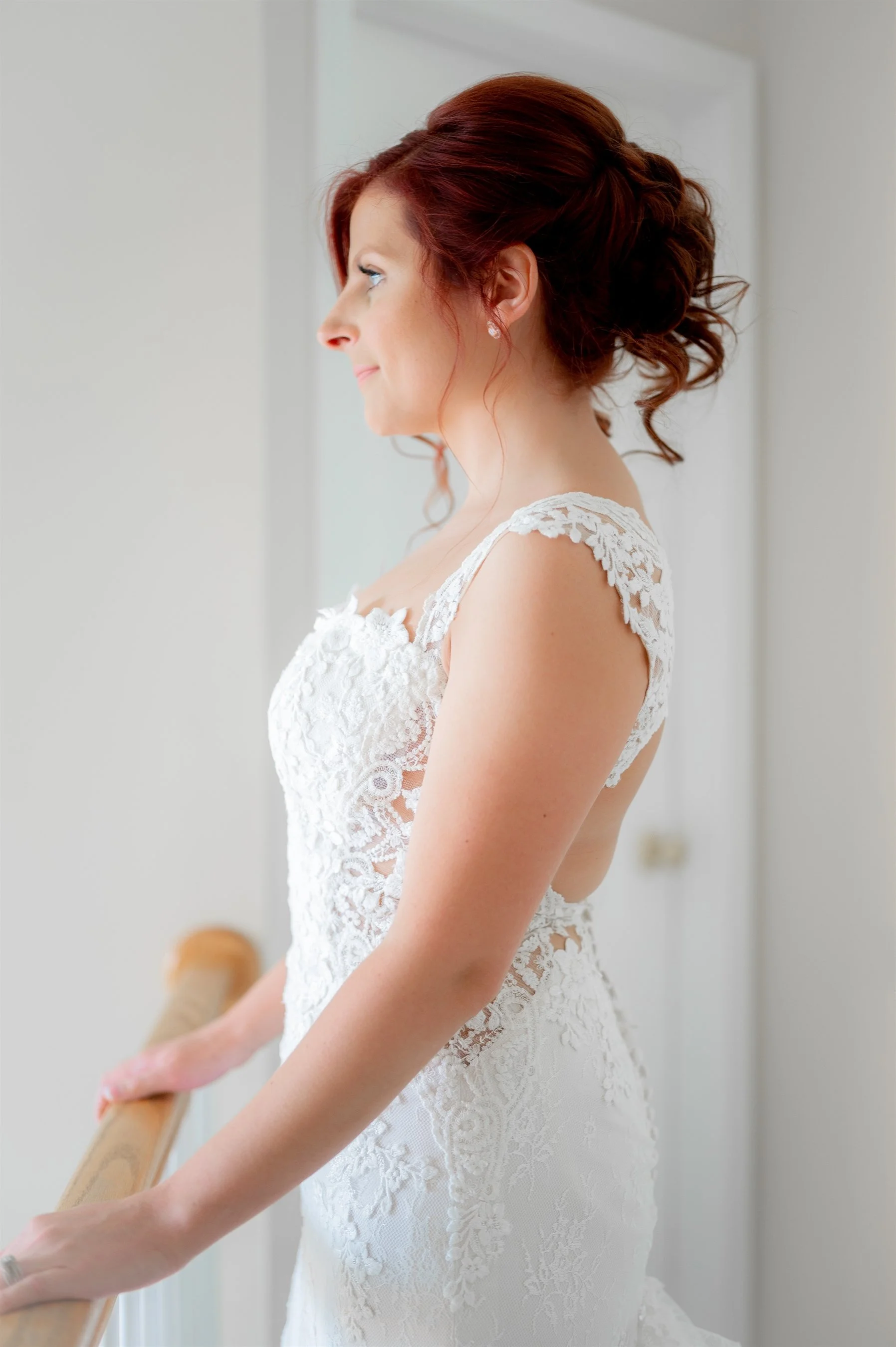 Bride in a lace wedding dress standing indoors, looking out of a window, with red hair styled in an elegant updo.