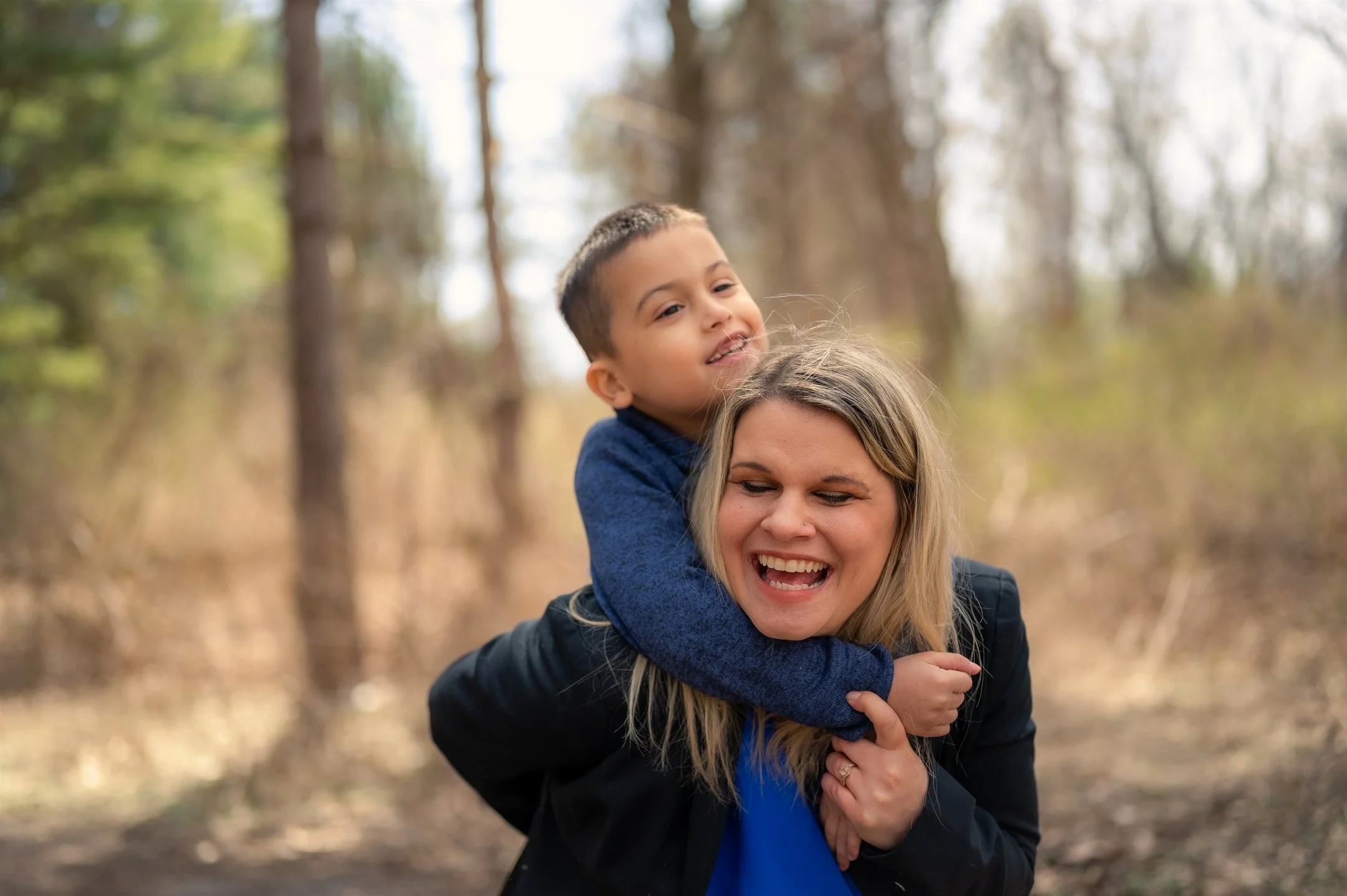 A woman smiling and giving a piggyback ride to a young boy in a wooded area.