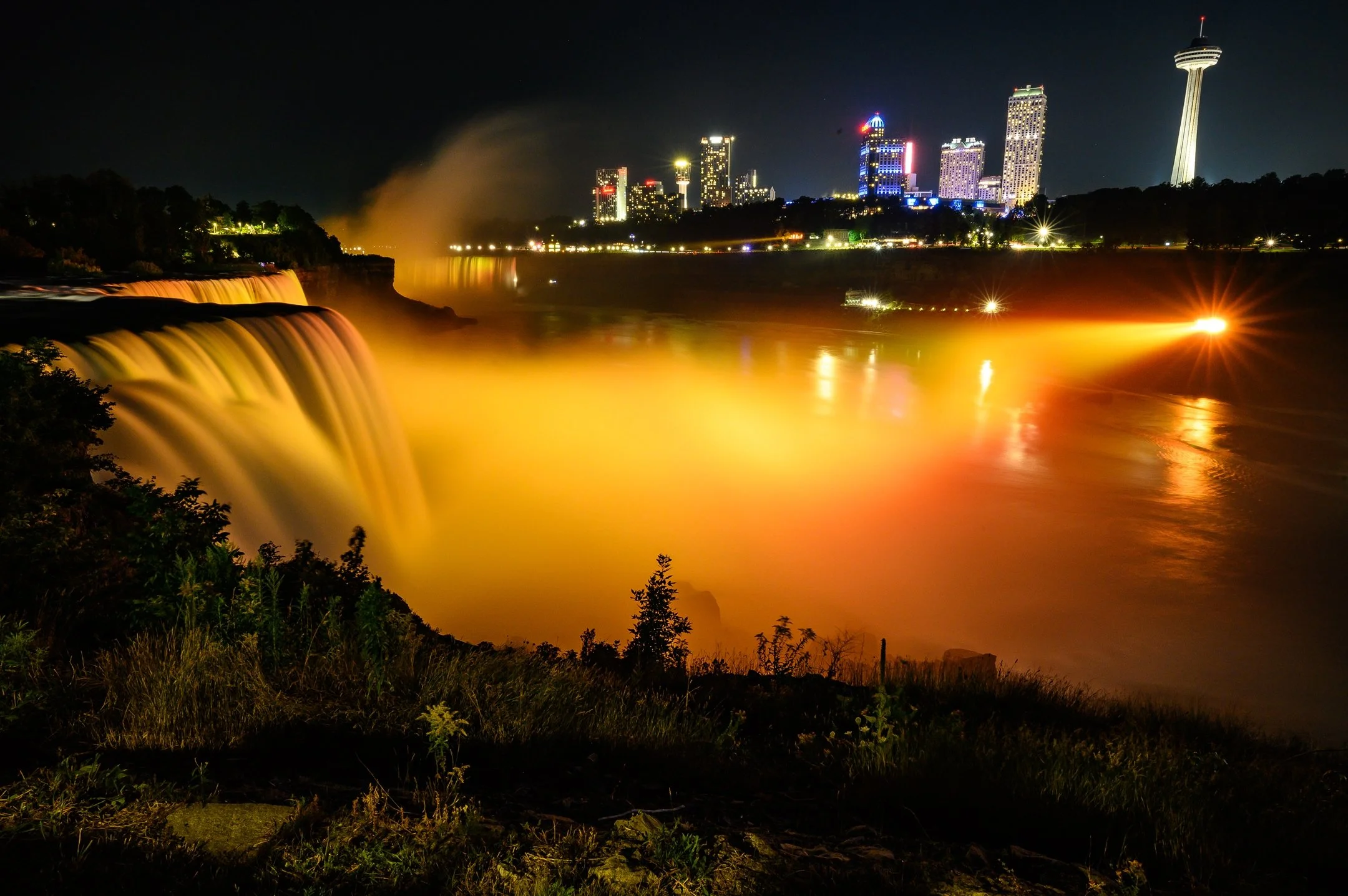 Night view of illuminated Niagara Falls with city skyline in the background, featuring colorful lights reflecting on the water.