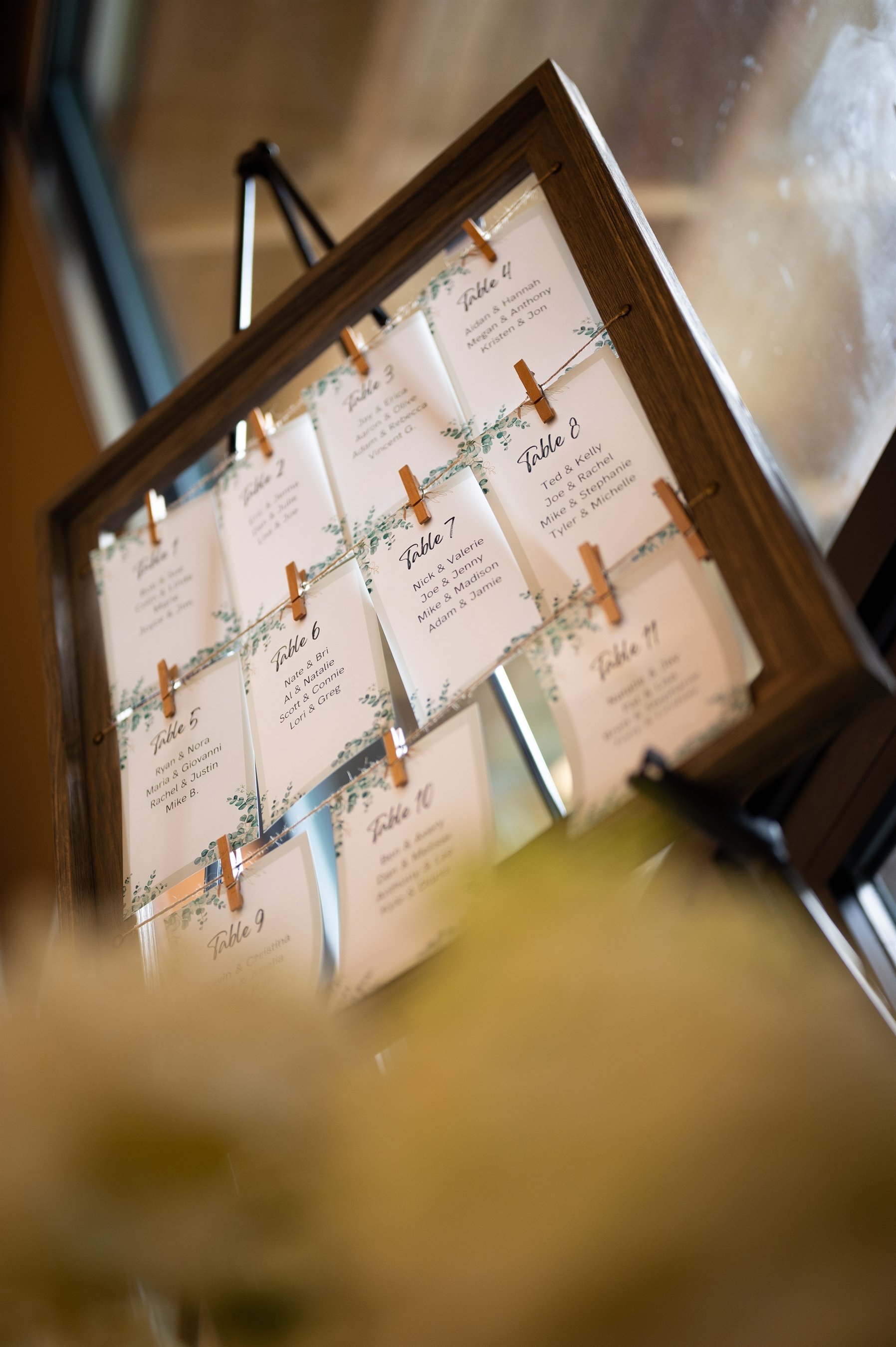 Wedding seating chart display with table assignments on a wooden frame, using clothespins to hold cards.