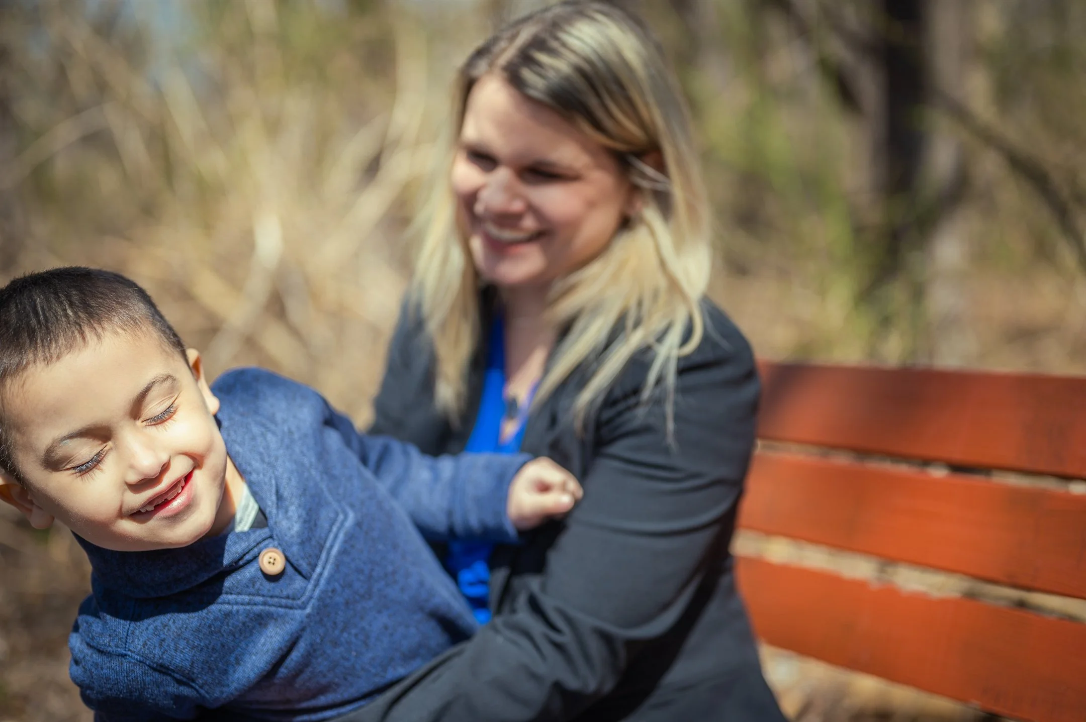 A woman and a young boy smiling and sitting on a wooden bench outdoors.
