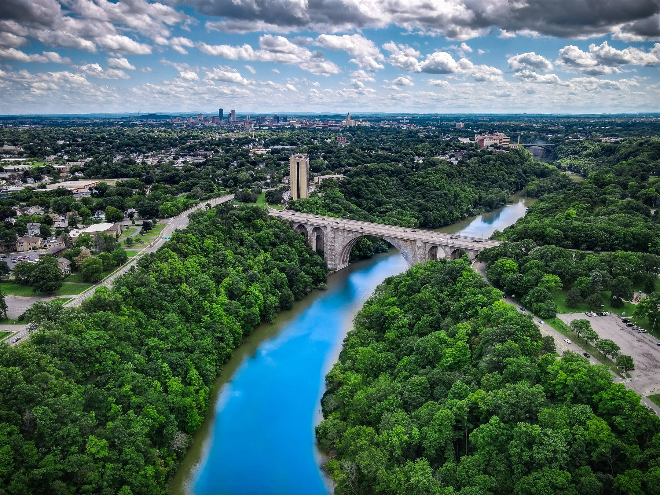 Aerial view of a lush green landscape featuring a river with a bridge crossing over it, surrounded by dense forests and a city skyline in the background under a partly cloudy sky.