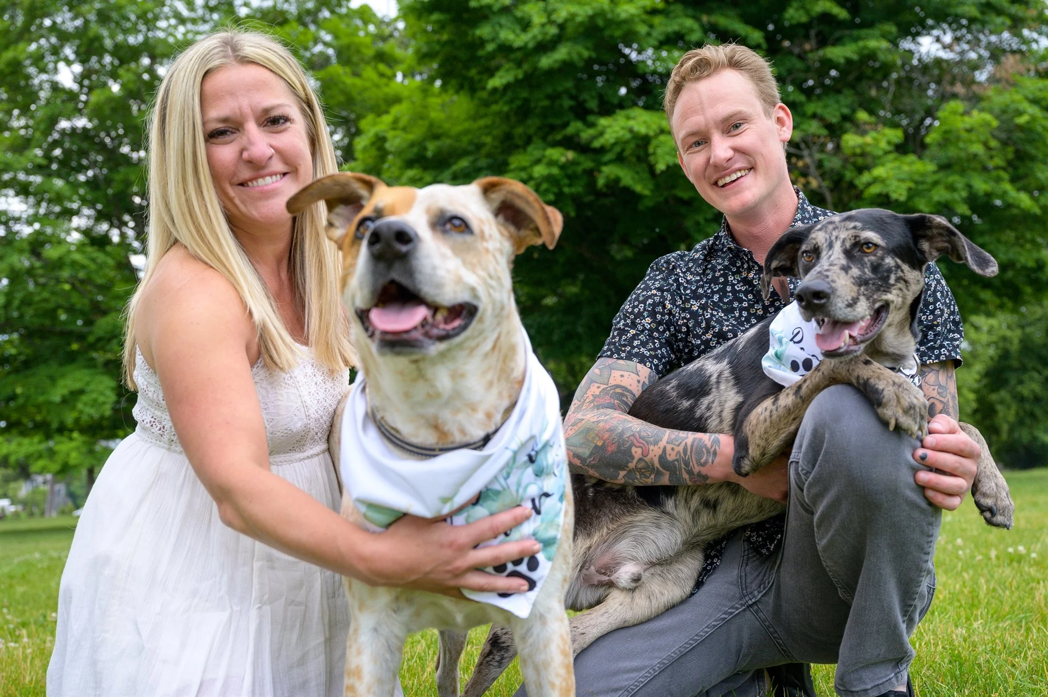 A woman and man sitting on the grass with two dogs, smiling. The woman has long blonde hair and wears a white dress, holding a dog with a spotted coat and a white bandana. The man has tattoos, short hair, and a patterned shirt, holding a brindle dog 