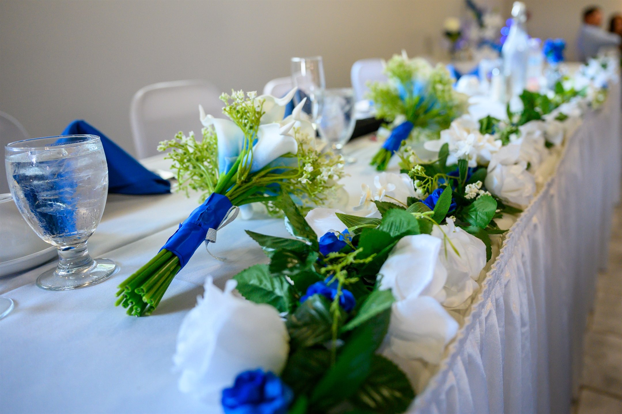 A wedding reception table decorated with a white tablecloth, blue napkins, white and blue floral arrangements, and wine glasses filled with water.