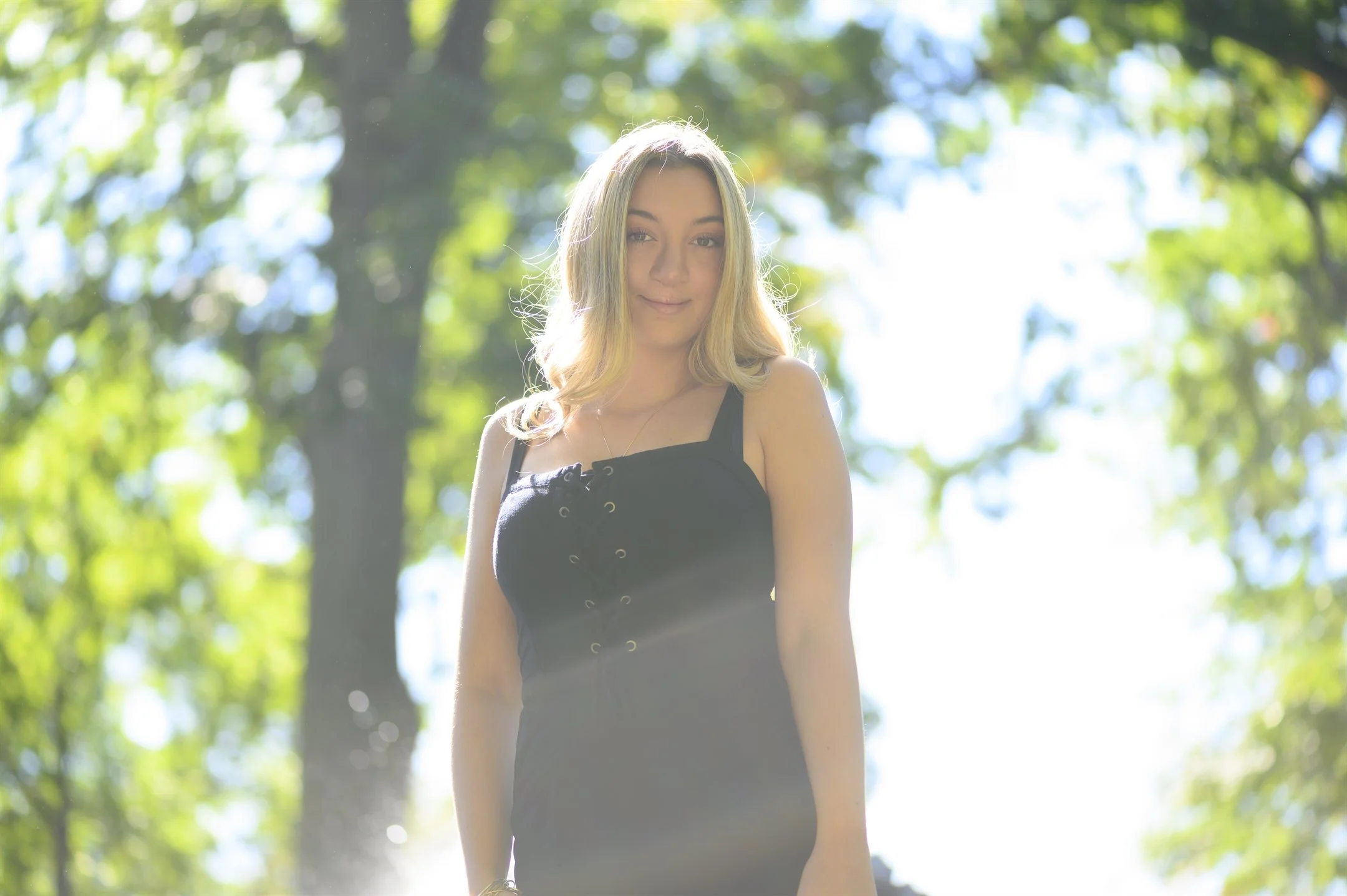 Woman in black dress standing outdoors with sunlight filtering through trees.