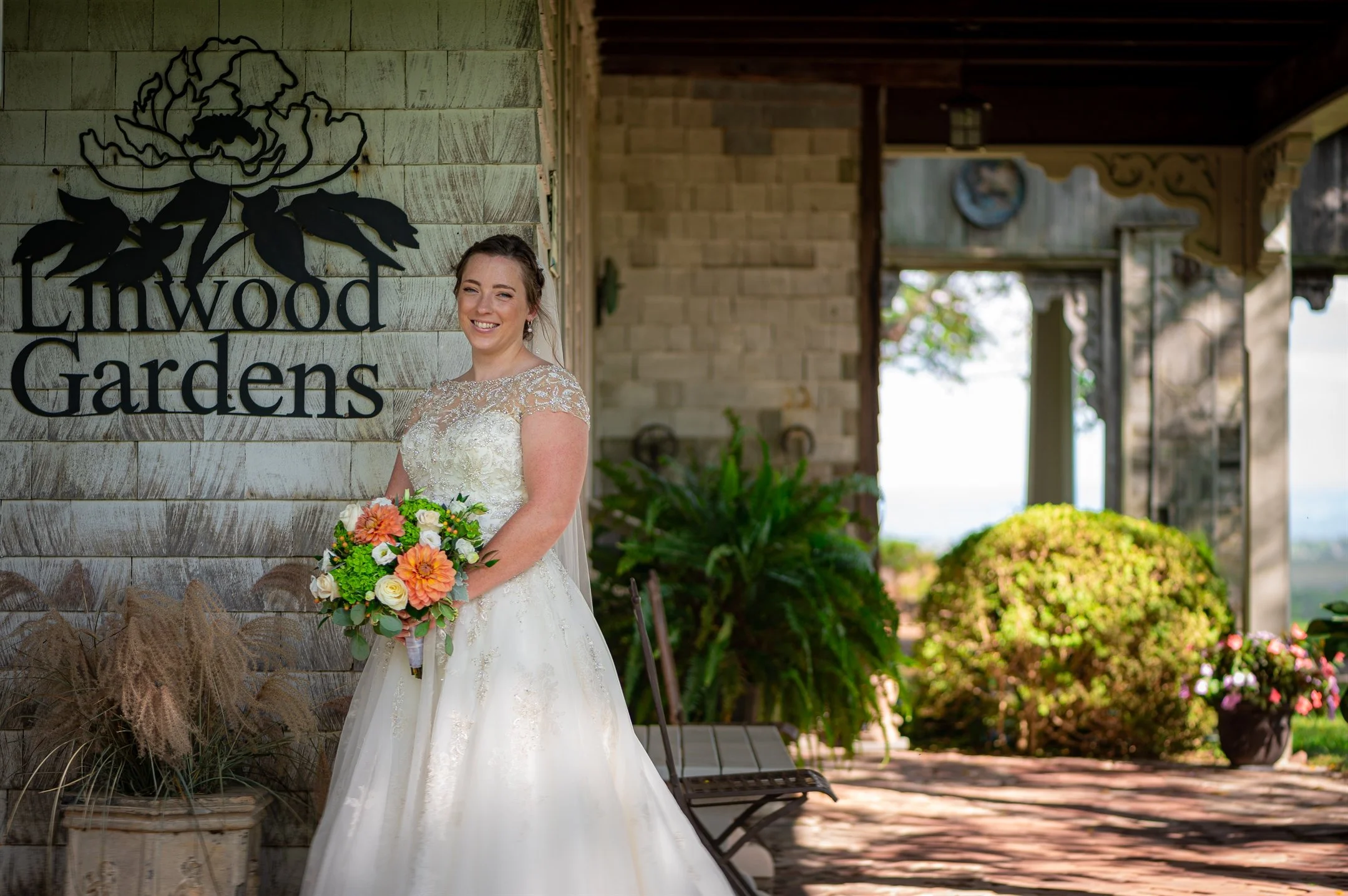 Bride in a white dress holding a bouquet at Linwood Gardens, with a rustic building and plants in the background.