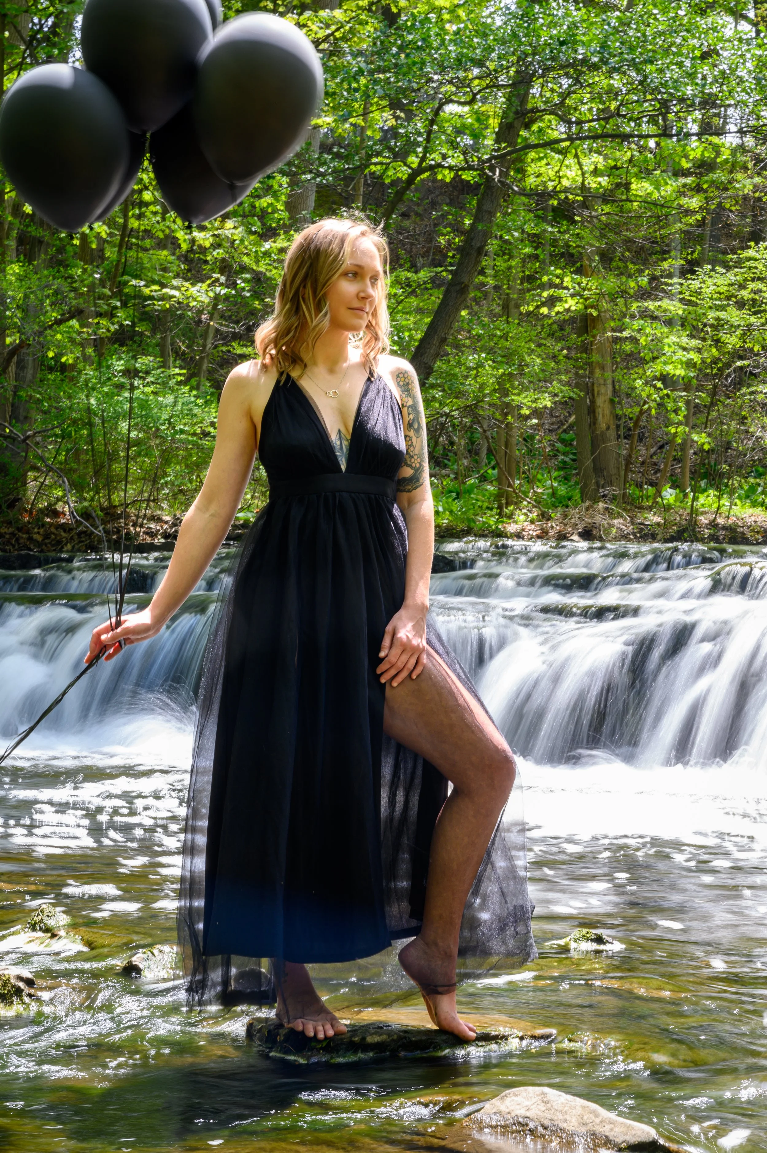 Woman in a black dress with black balloons near a waterfall in a forest.