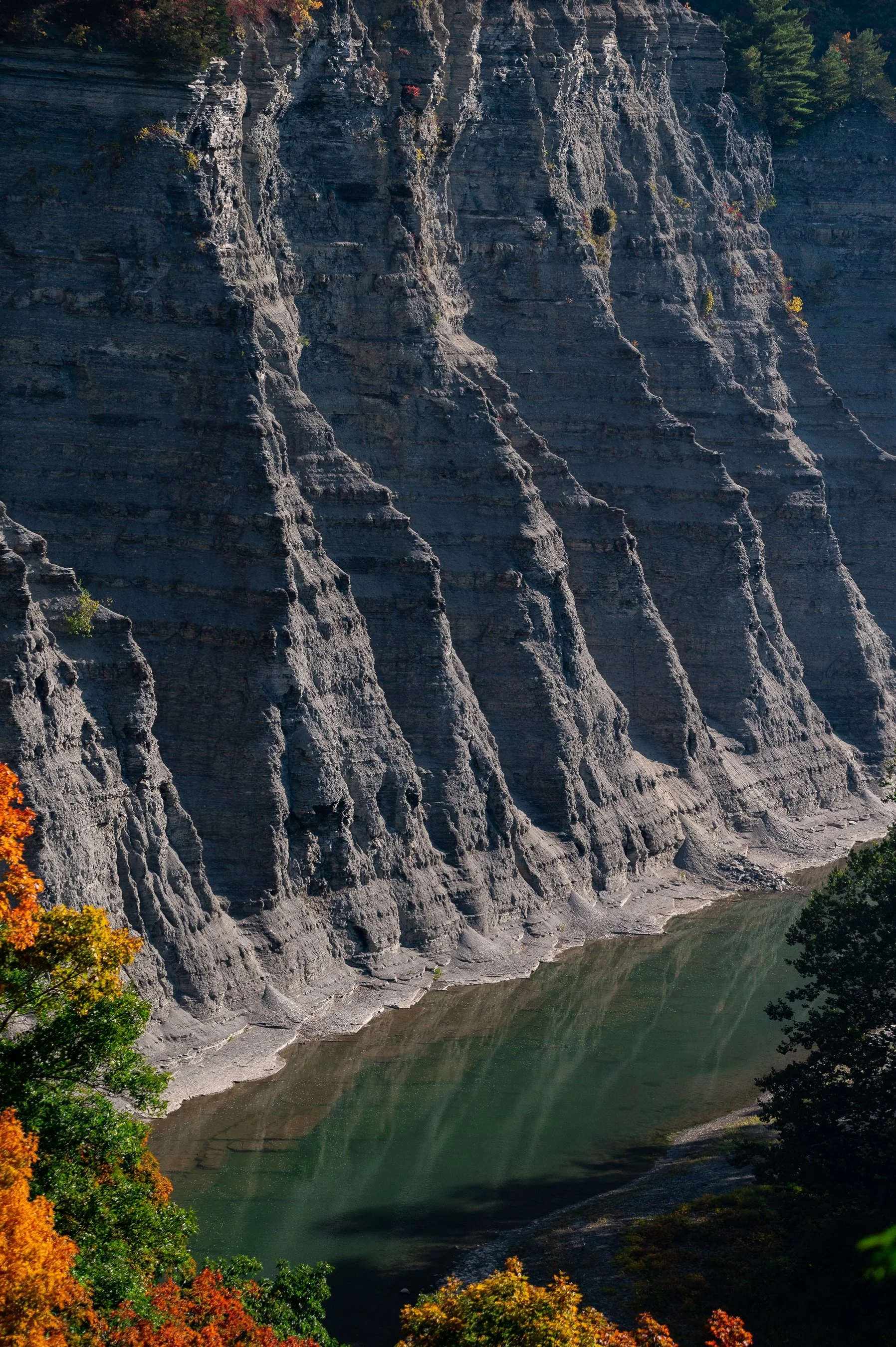 Tall rock cliff with layered formations next to a calm green river, surrounded by autumn-colored trees.