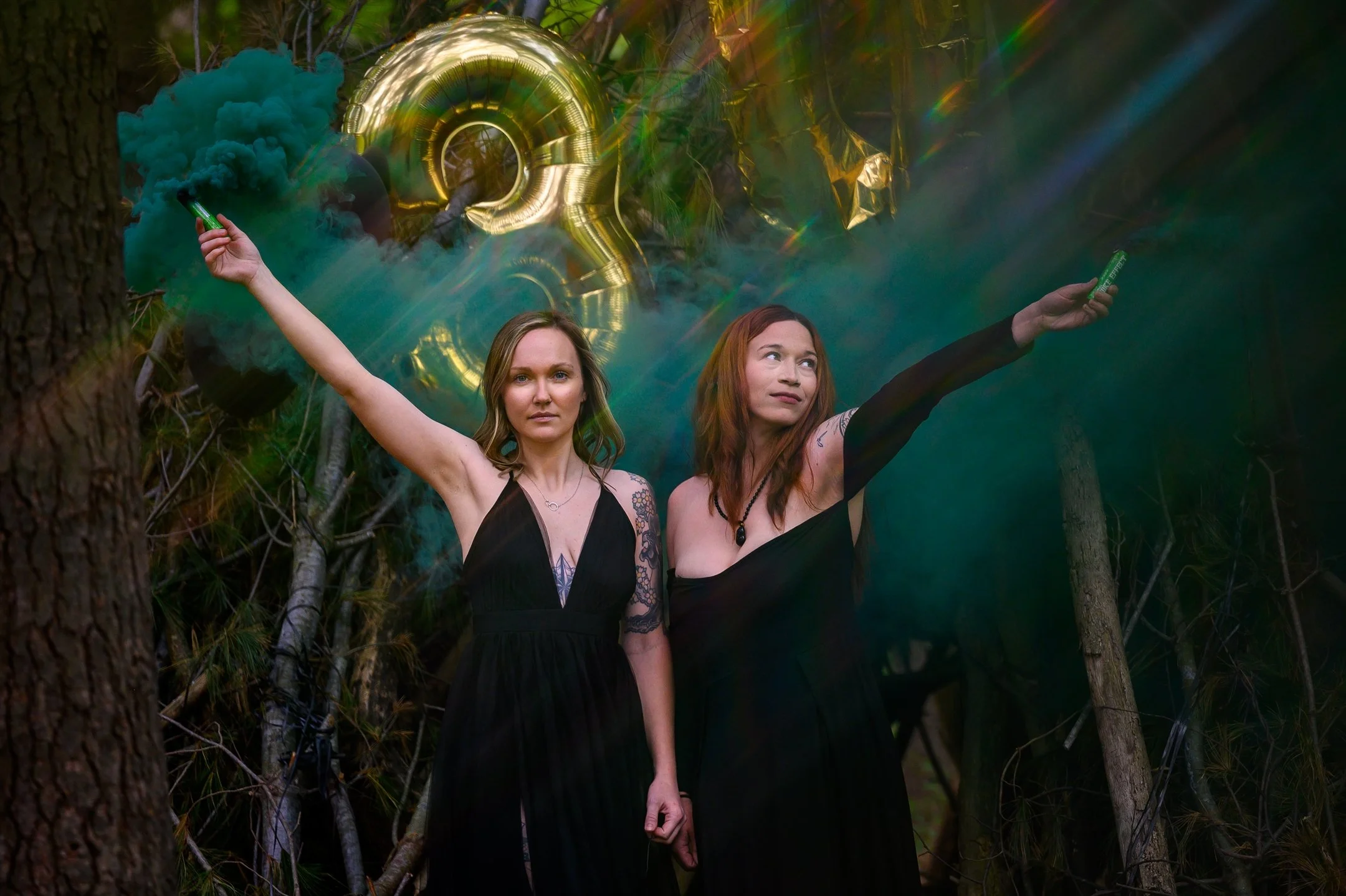 Two women in black dresses holding smoke sticks near a large gold number 3 balloon in a wooded area.