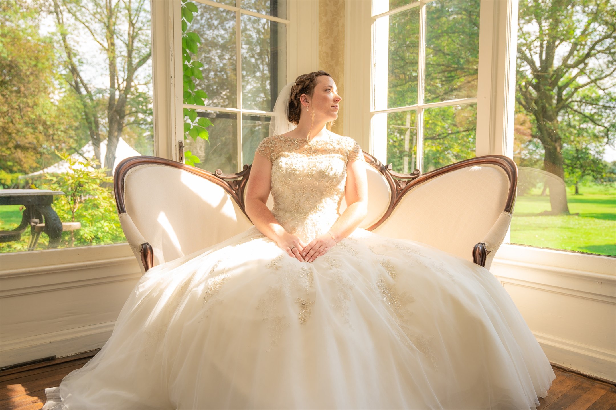 Bride sitting on antique sofa in elegant wedding dress near large windows with scenic garden view.