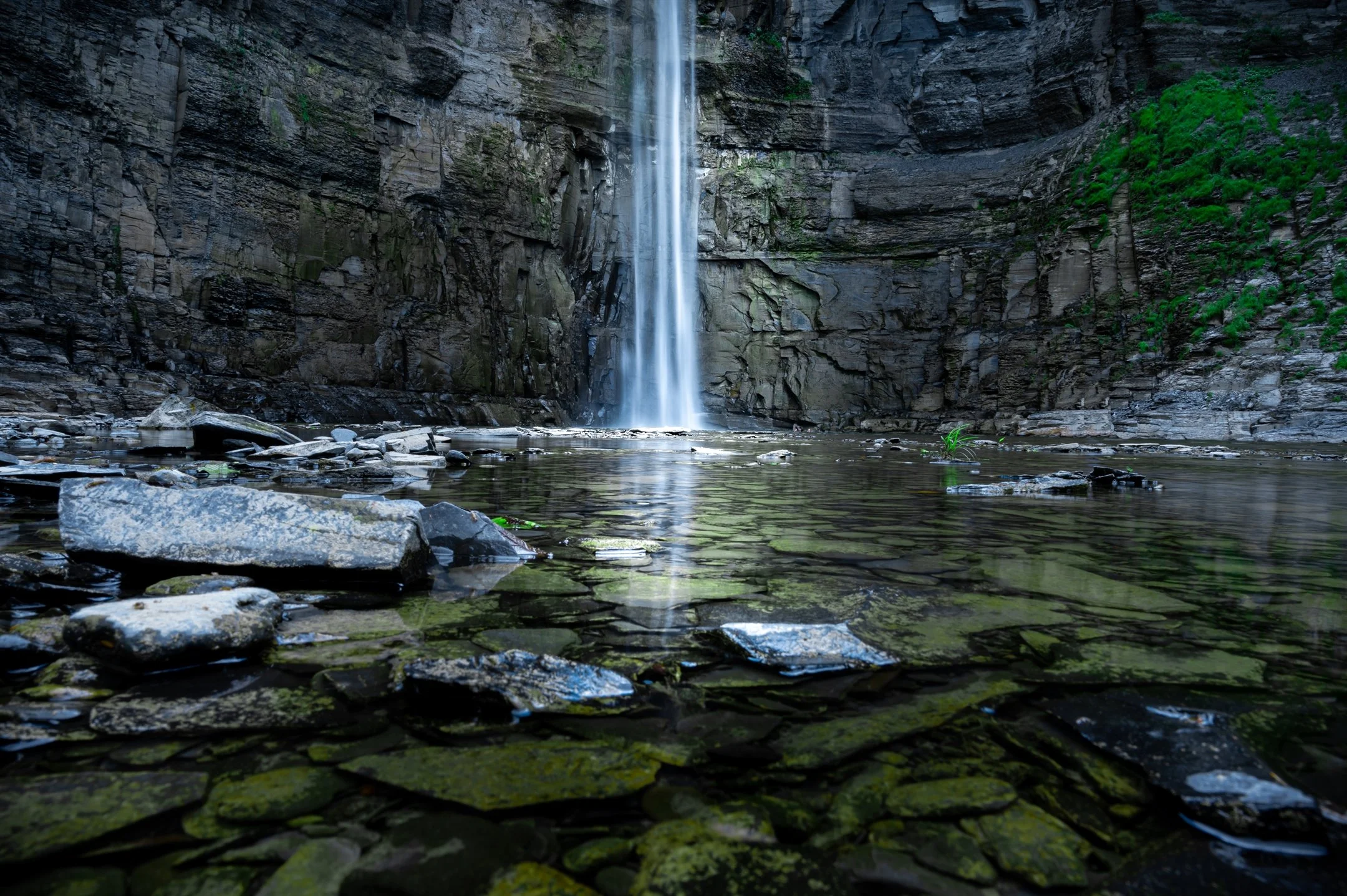 Waterfall cascading down a rocky cliff into a shallow pool with scattered stones, surrounded by green moss and foliage.