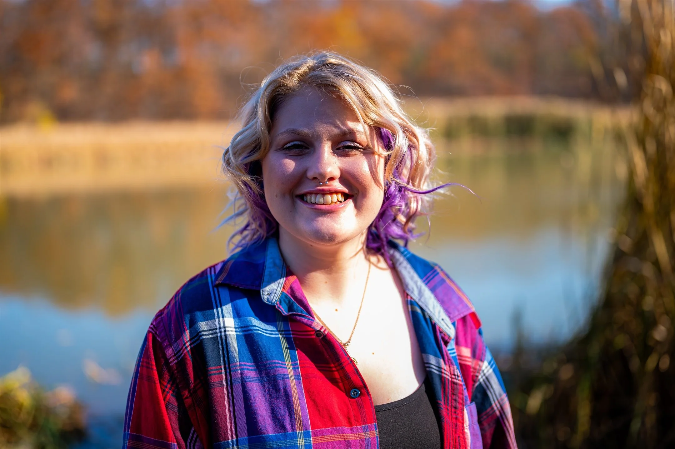Woman with curly blonde and purple hair wearing a plaid shirt, smiling outdoors with a lake and autumn trees in the background.