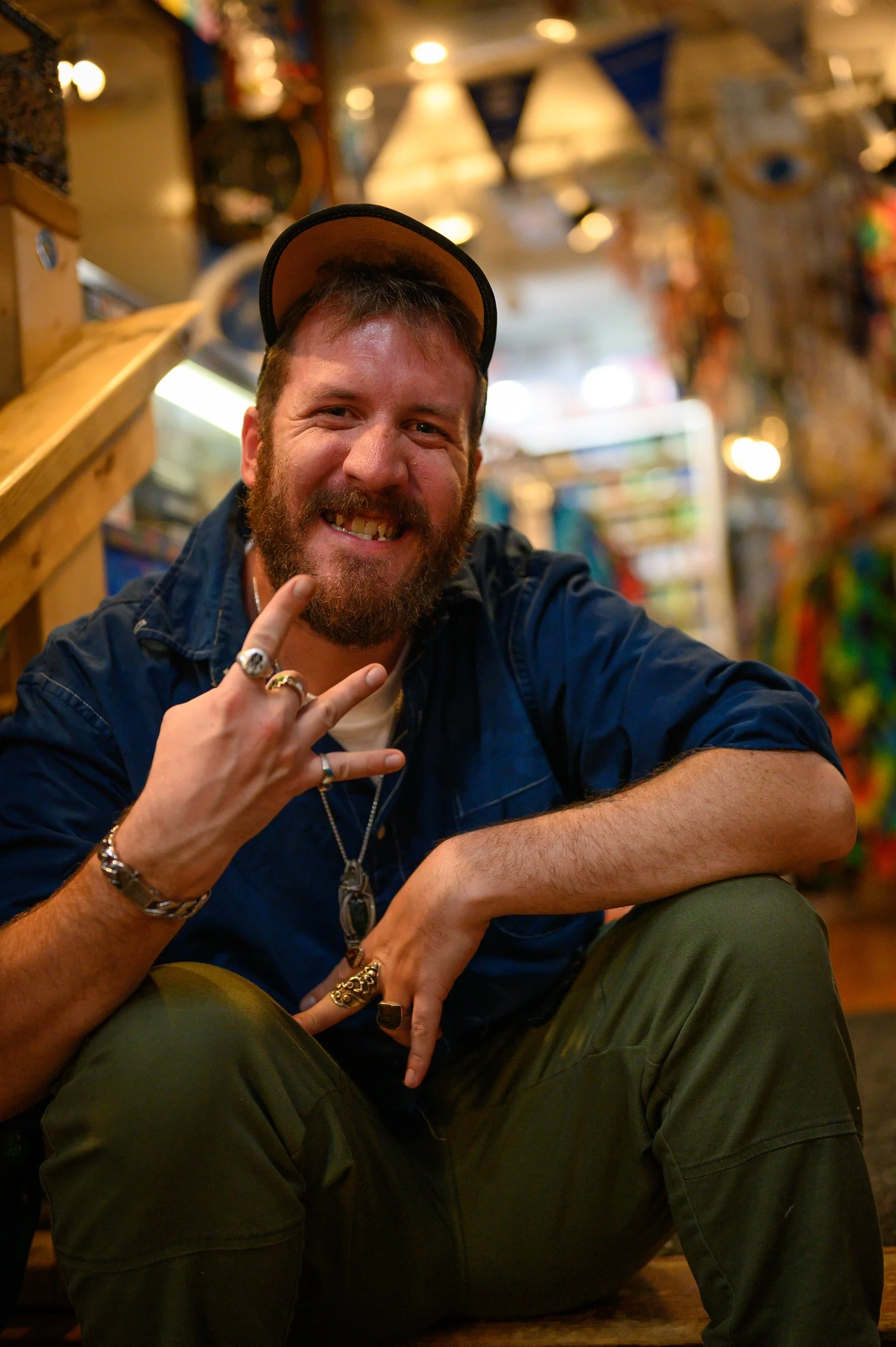 Man sitting indoors, wearing a cap and jewelry, smiling, making hand gesture.