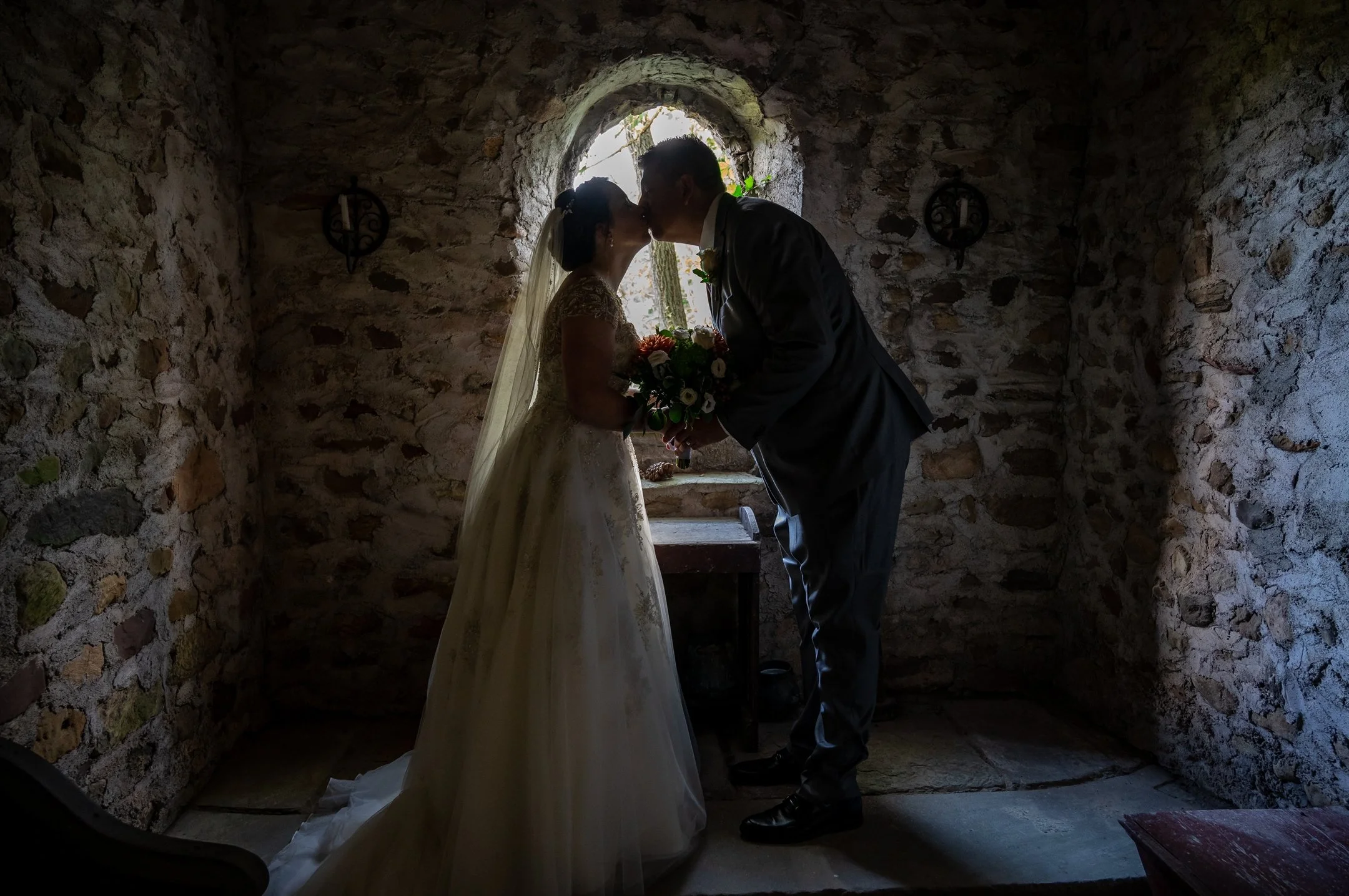 Silhouette of a bride and groom kissing in front of a small stone window, with the bride holding a bouquet.