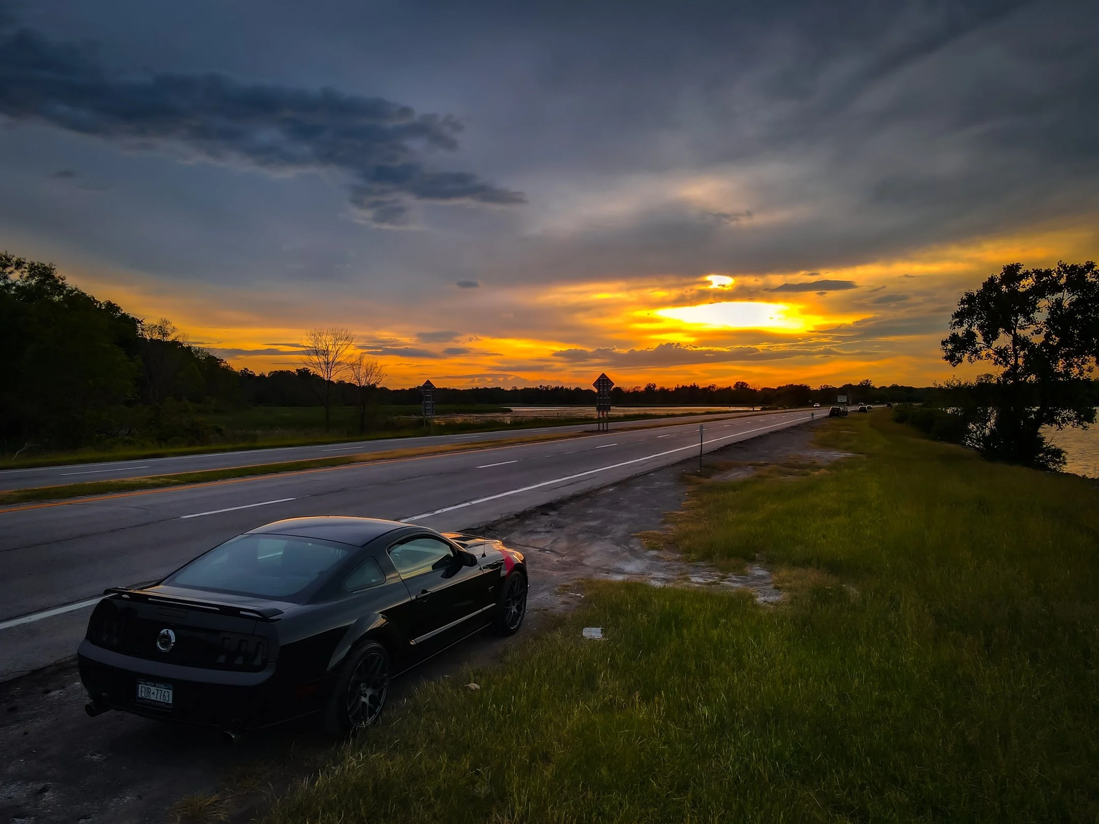 Black sports car parked beside a road with a scenic sunset and cloudy sky.