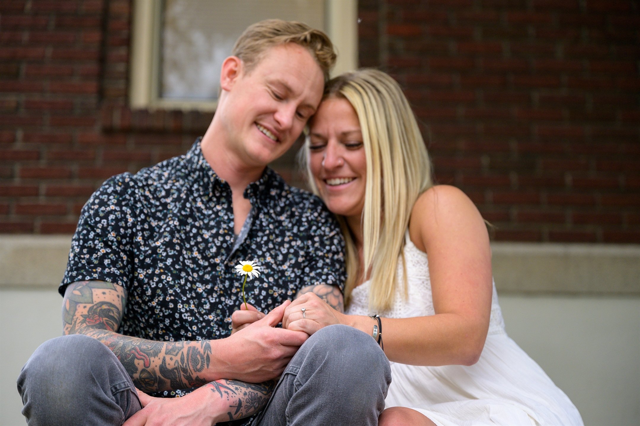 Couple sitting together holding a daisy, smiling and leaning on each other, with a brick wall in the background.