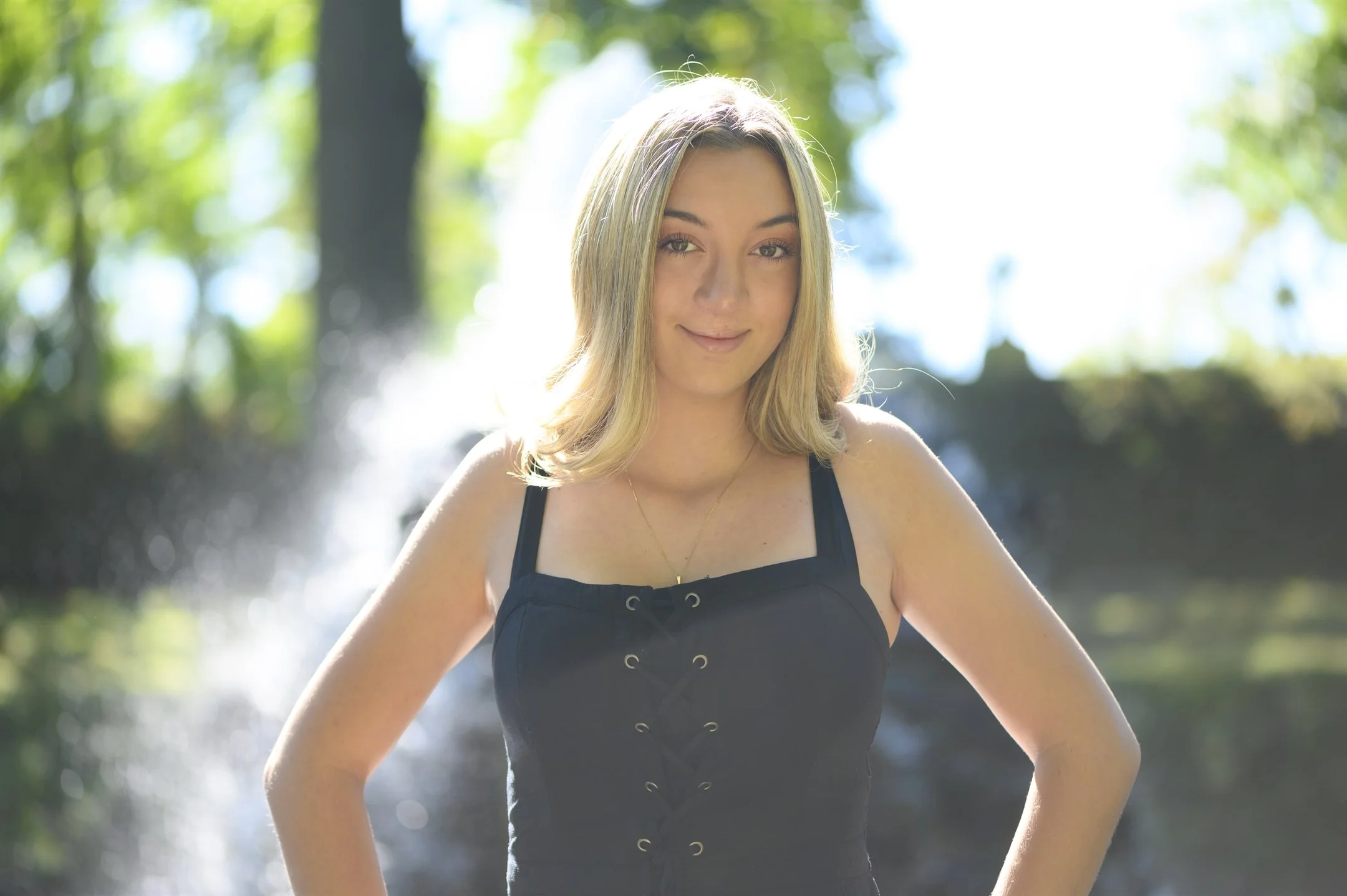 Woman with blonde hair wearing a black top, standing outdoors with trees and sunlight in the background.