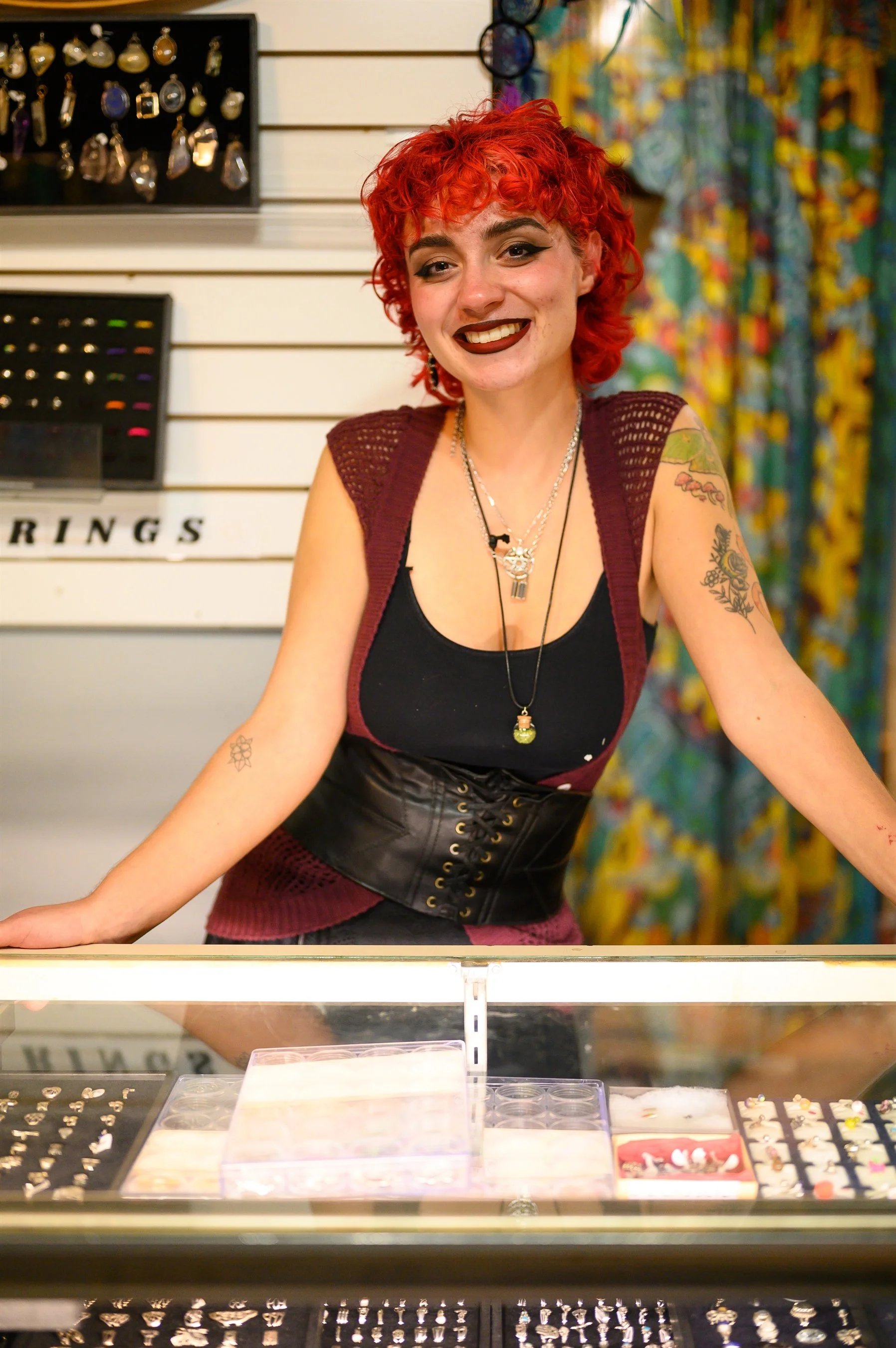Person with red curly hair standing behind a jewelry display case, wearing a black top, purple vest, and black lace-up corset, with earrings on display in the background.