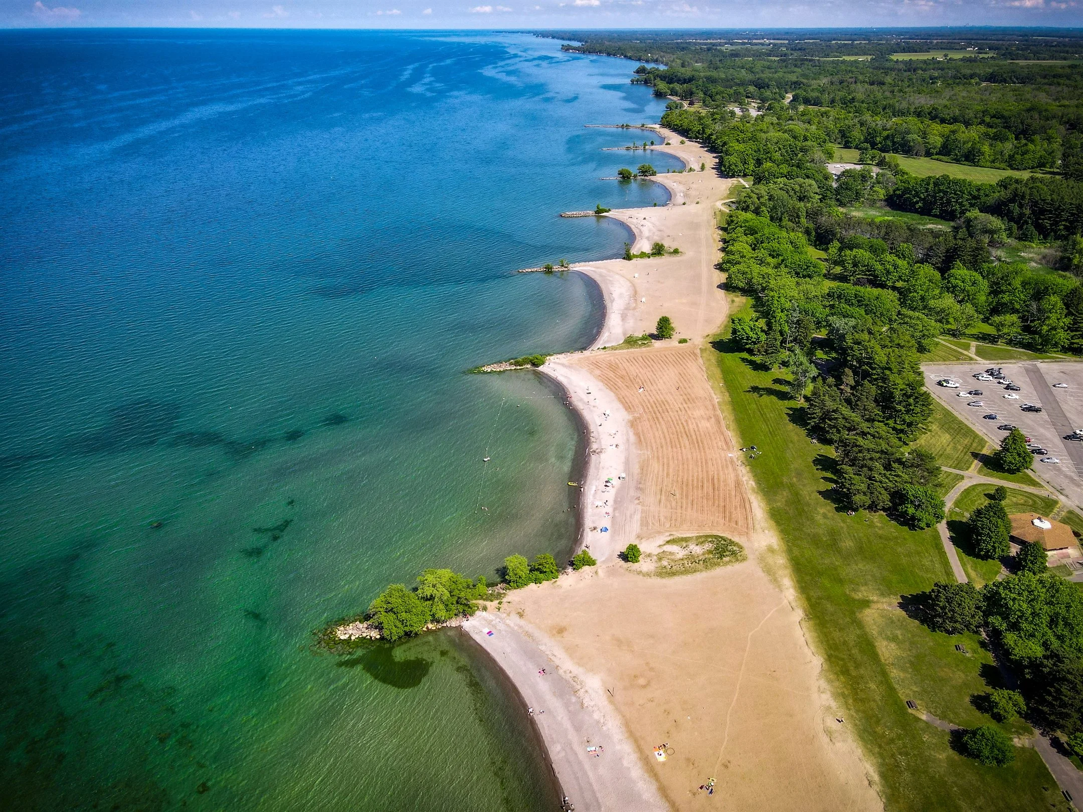 Aerial view of a large, sandy beach adjacent to a calm, clear blue lake. The beach is partly bordered by lush green trees and a parking lot to the right. There are small groups of people scattered across the beach. The shoreline curves gently along t