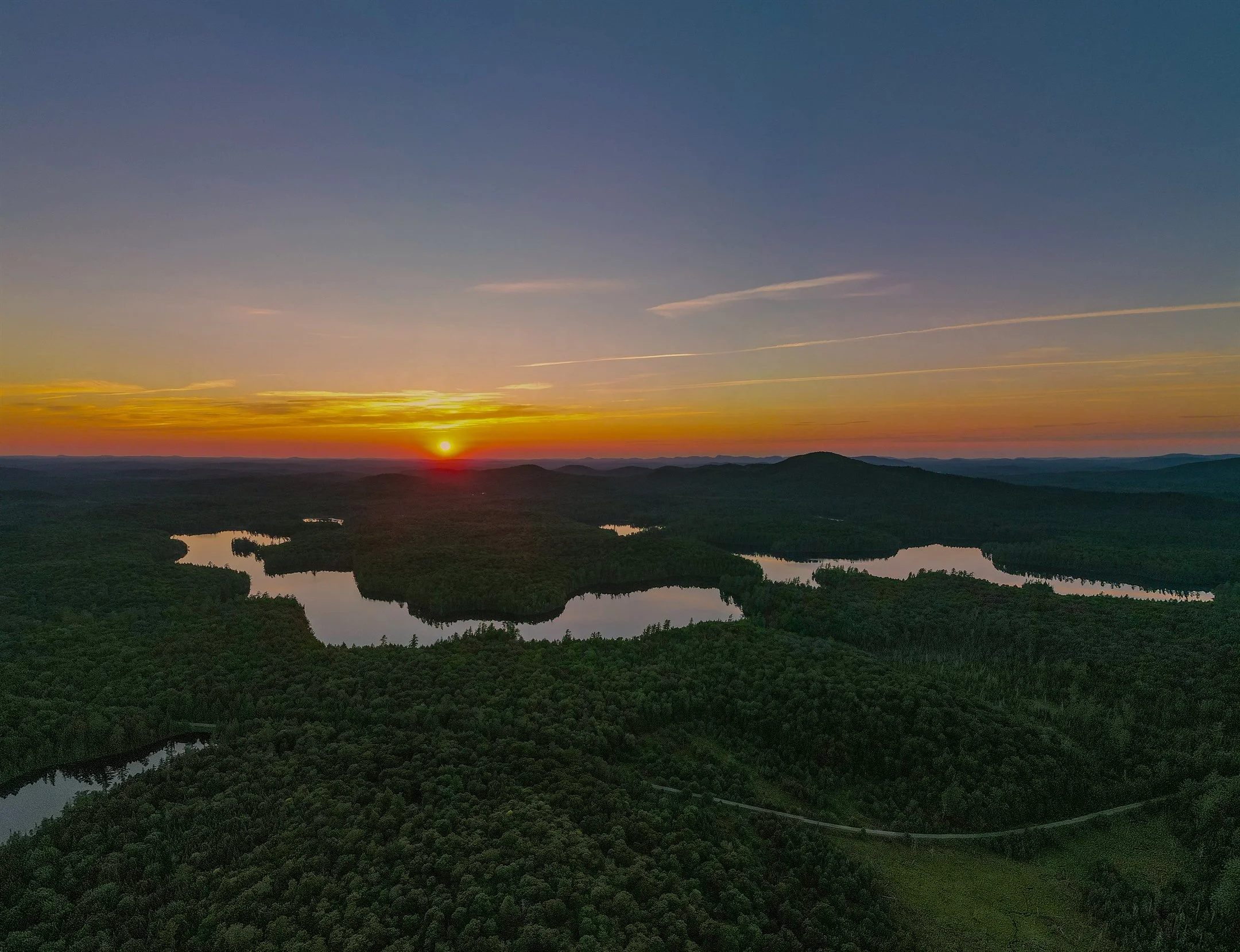 Scenic sunset view over a forest with lakes and distant mountains.