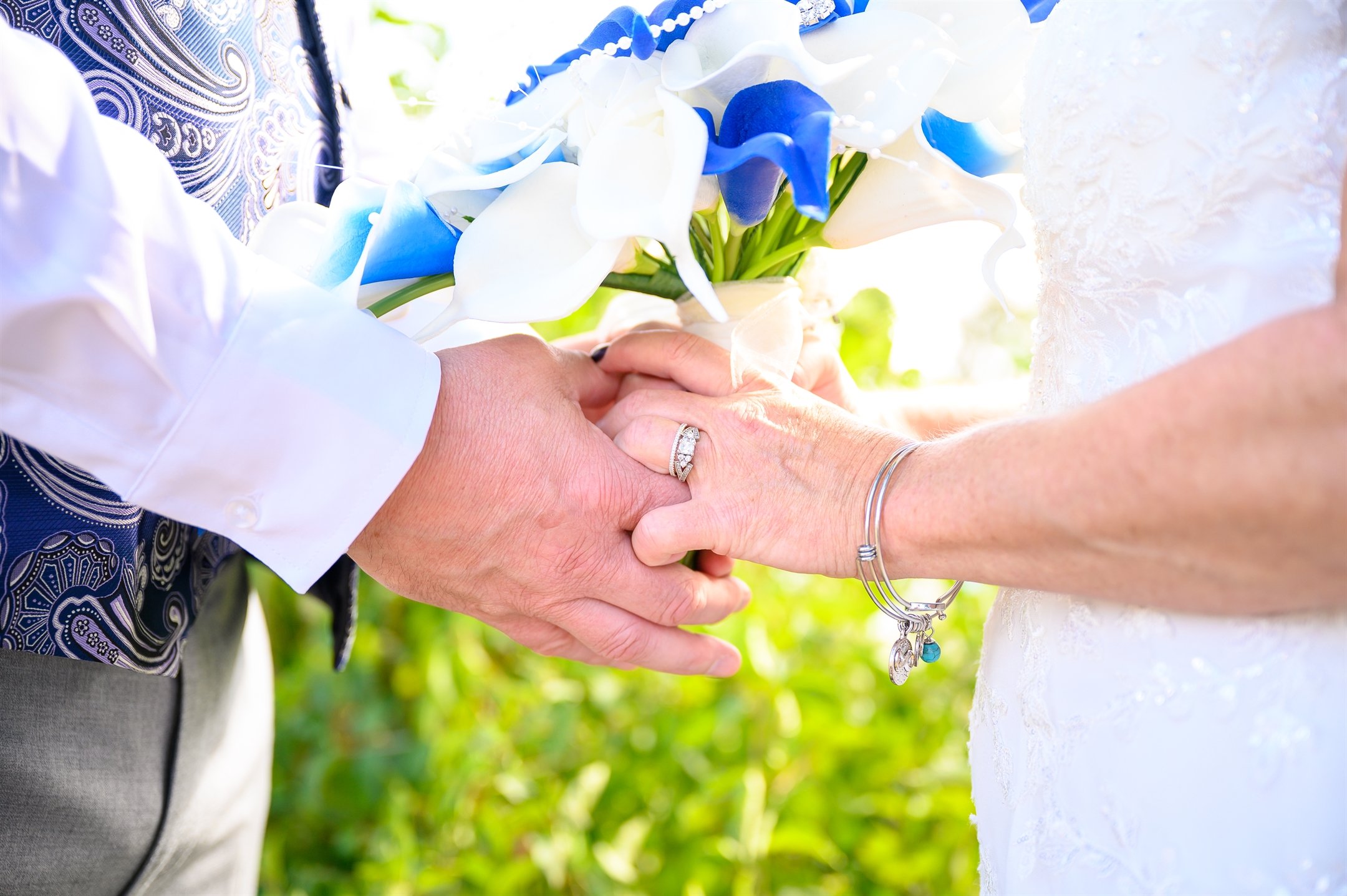Close-up of a bride and groom holding hands with a bouquet of blue and white flowers, showing their wedding rings.