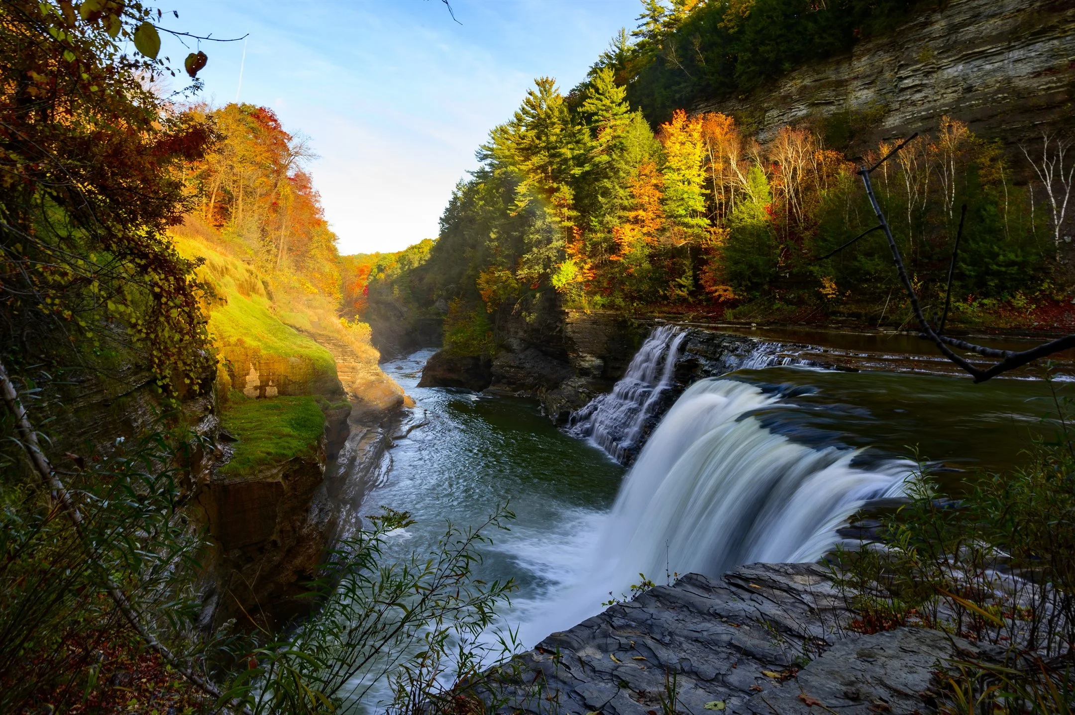 Scenic waterfall surrounded by autumn foliage in a forested gorge, featuring sunlight illuminating colorful trees and rocky cliffs.