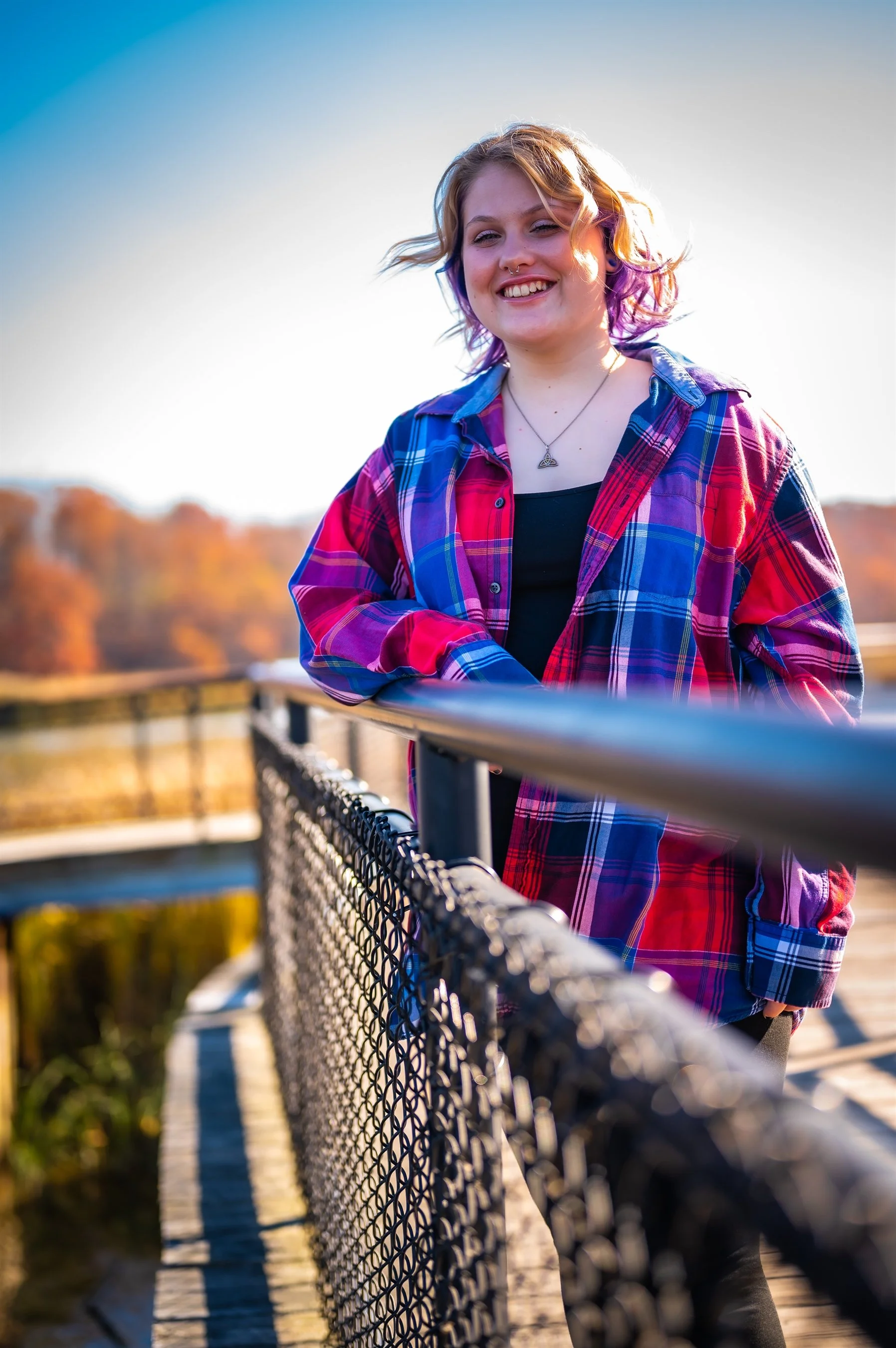 A person wearing a red and blue plaid shirt leans on a chain-link fence on a wooden boardwalk, with autumn trees in the background.