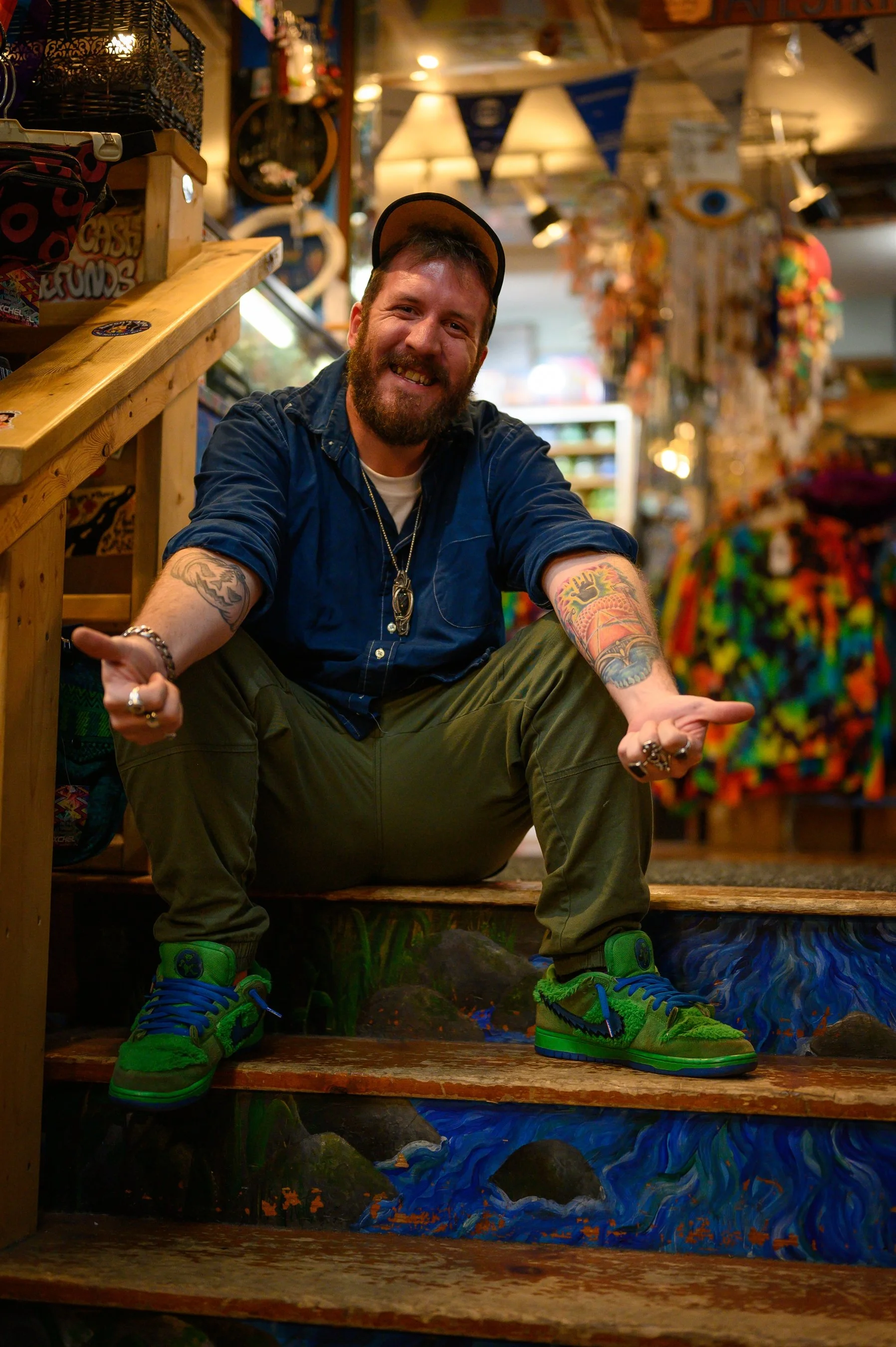 A man sitting on brightly painted wooden stairs in a colorful store, smiling. He wears a blue shirt, khaki pants, green sneakers, and a cap, and has tattoos on his arms. The background is filled with various vibrant items.