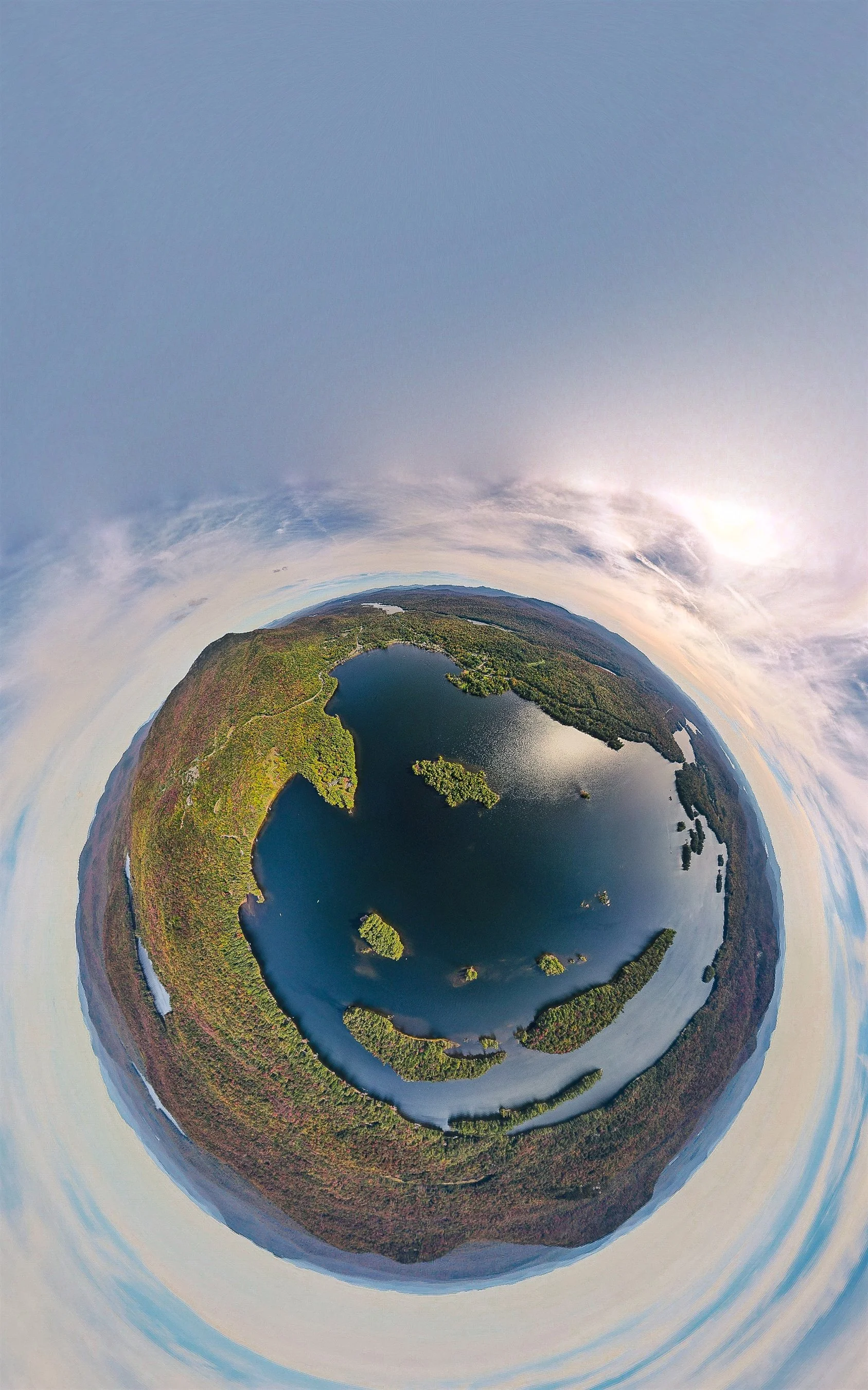 Aerial tiny planet view of a lake surrounded by forests and clouds.