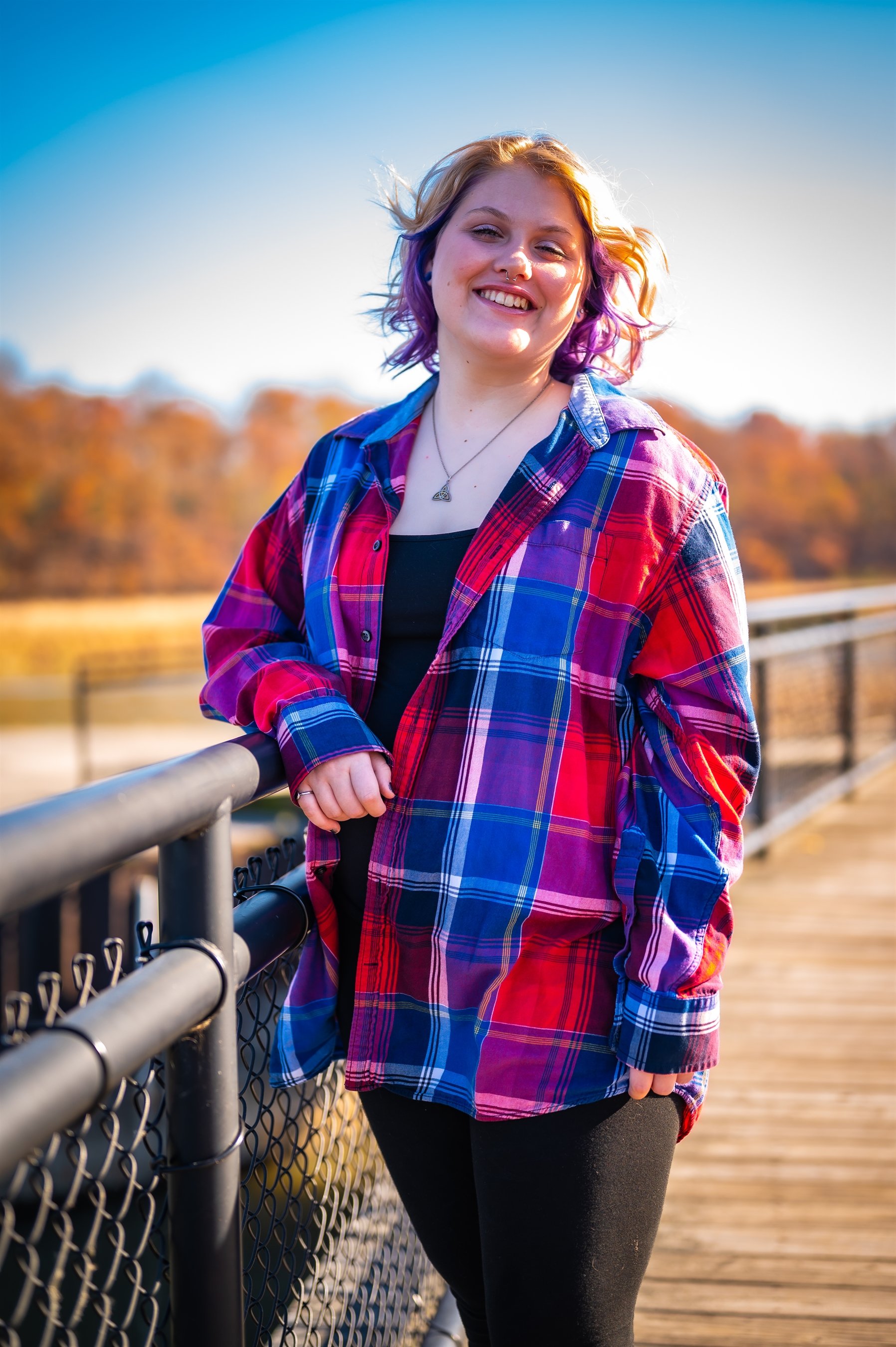 Person with purple hair and plaid shirt smiling near a fence on a sunny day.