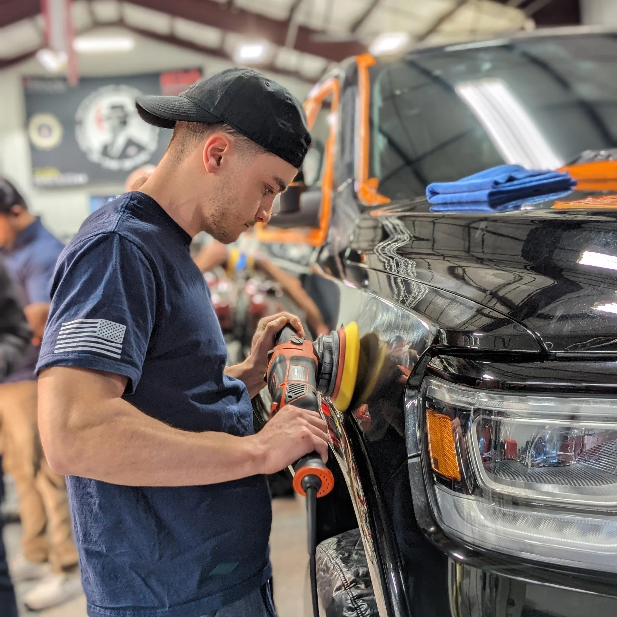 A young man polishing a black vehicle using a yellow buffer in a garage or auto detailing shop.