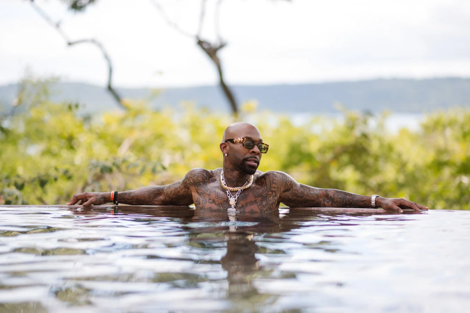 A tattooed man with sunglasses and jewelry relaxing in an outdoor pool with arms stretched out on the pool edge, with green trees and distant hills in the background.