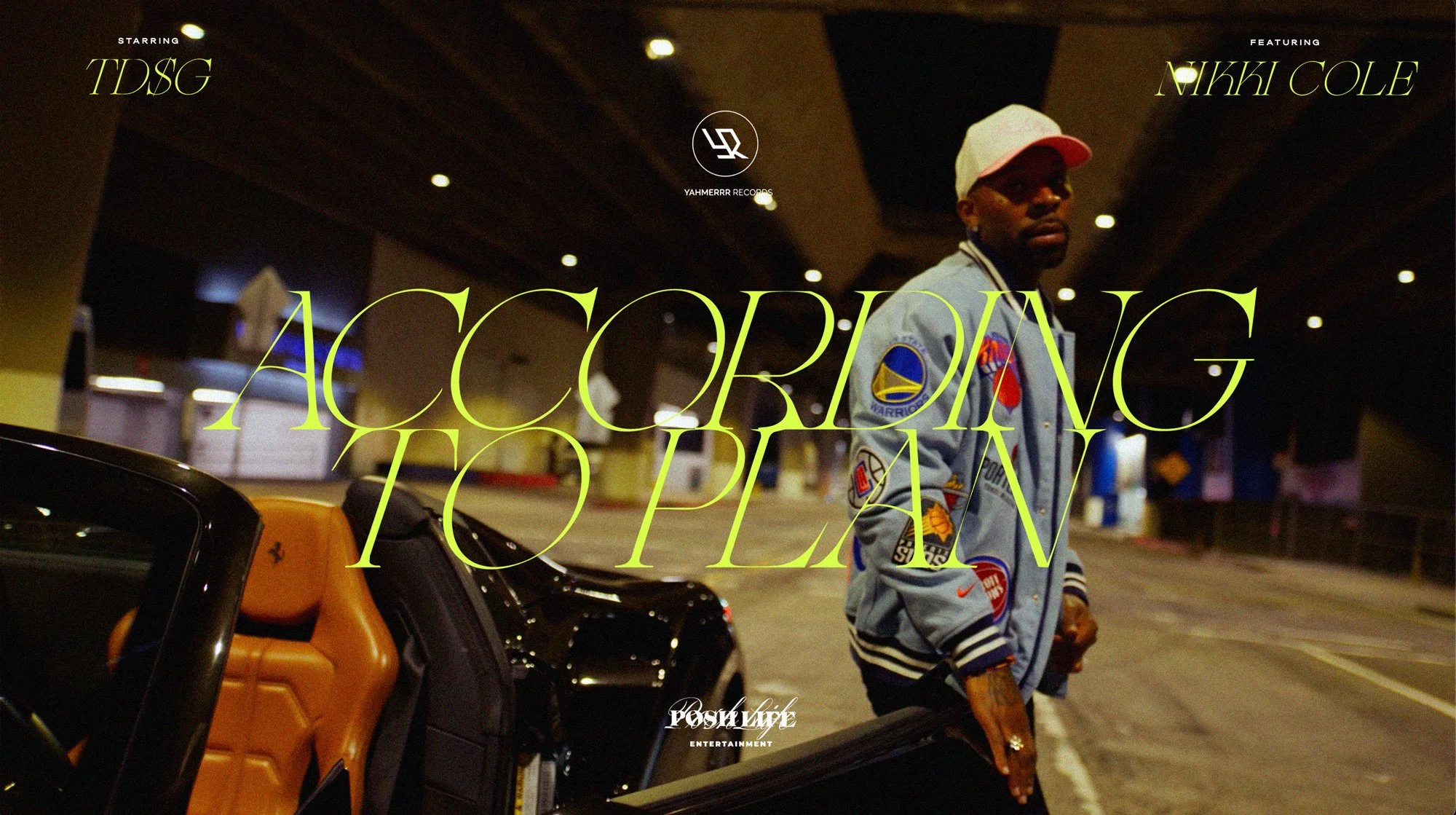 A man stands next to luggage and a go-kart under an indoor parking structure, wearing a light-colored jacket with patches and a white cap.