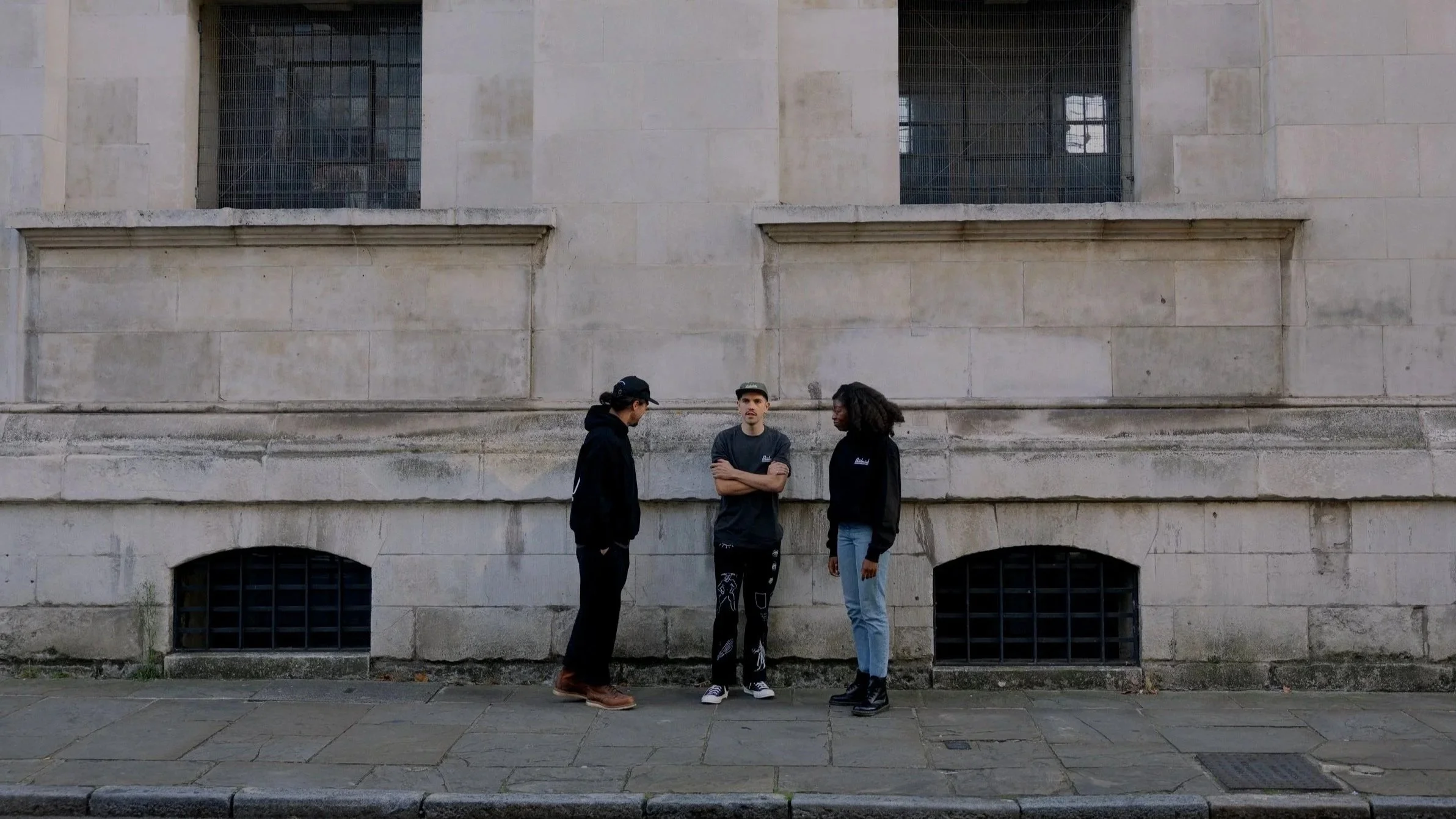 Three young people standing on a sidewalk against a large stone building wall, engaged in conversation. The person in the middle has arms crossed, wearing a cap, t-shirt, and patterned pants. The person on the left is in a black hoodie and cap, looking down. The person on the right wears a black hoodie and light blue jeans, looking at the person in the middle.