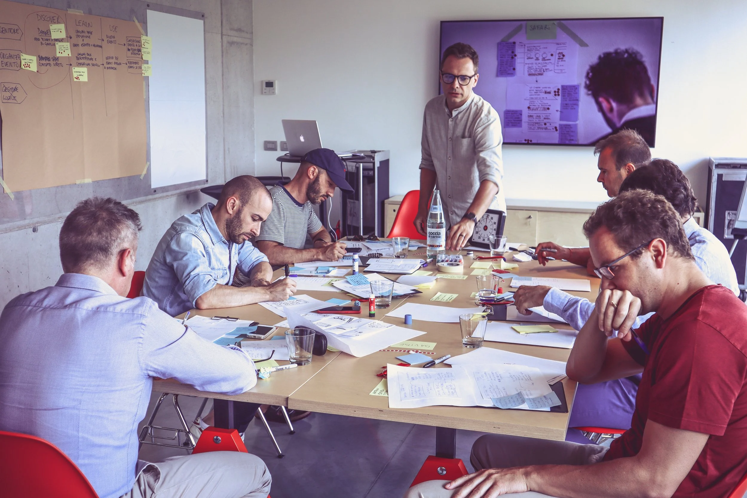 Un gruppo di persone sedute attorno a un tavolo durante una sessione di lavoro, con documenti, appunti e penne, mentre una persona si trova in piedi accanto al tavolo, che sembra spiegare o presentare qualcosa.