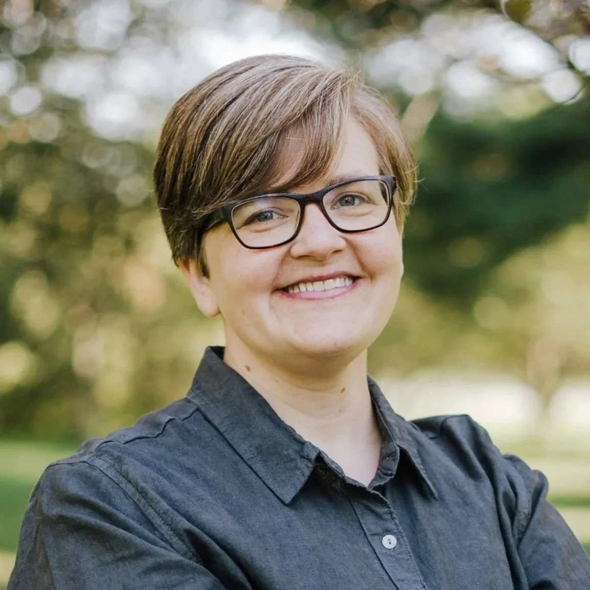 Smiling woman in glasses with short hair, wearing a dark button-up shirt, standing outdoors with a blurred background of trees and grass.